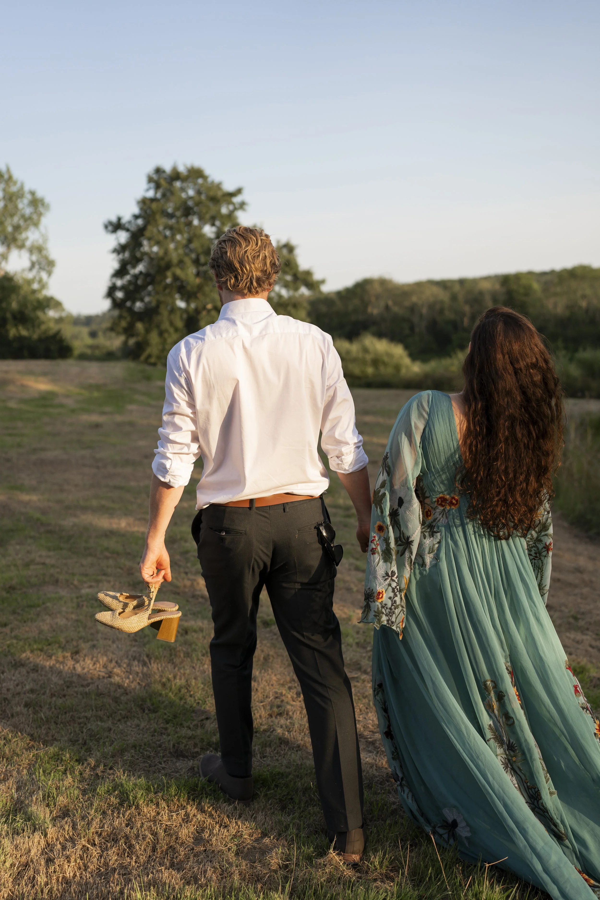 A man and woman walk away from the camera in an open field during sunset, holding hands. The man is dressed in a white shirt and black pants, carrying a straw hat and sandals. The woman is in a flowing green dress with floral patterns and a matching cover-up.