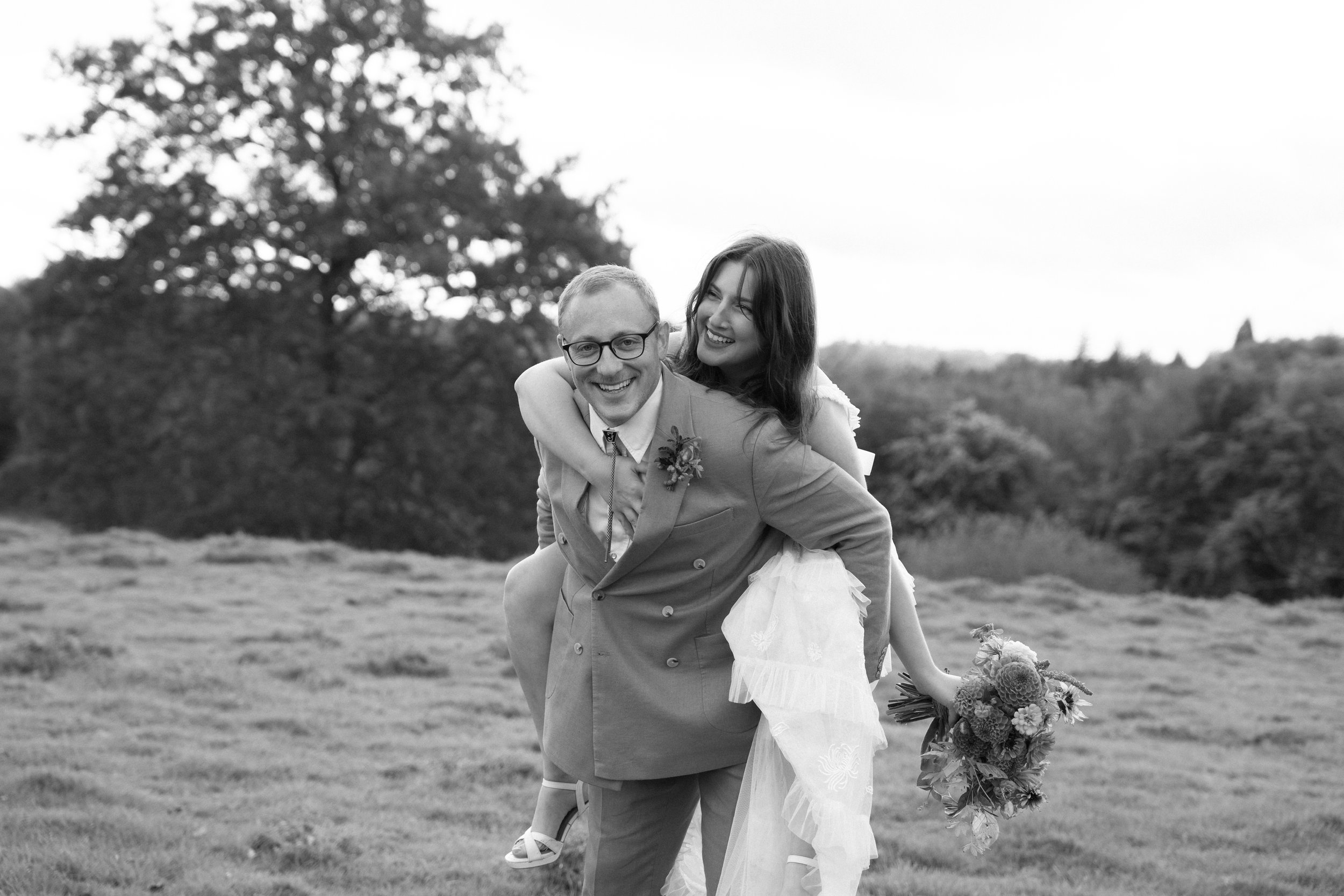 A smiling bride and groom in wedding attire outdoors, with the groom carrying the bride on his back, holding a bouquet of flowers, trees in the background, in black and white.