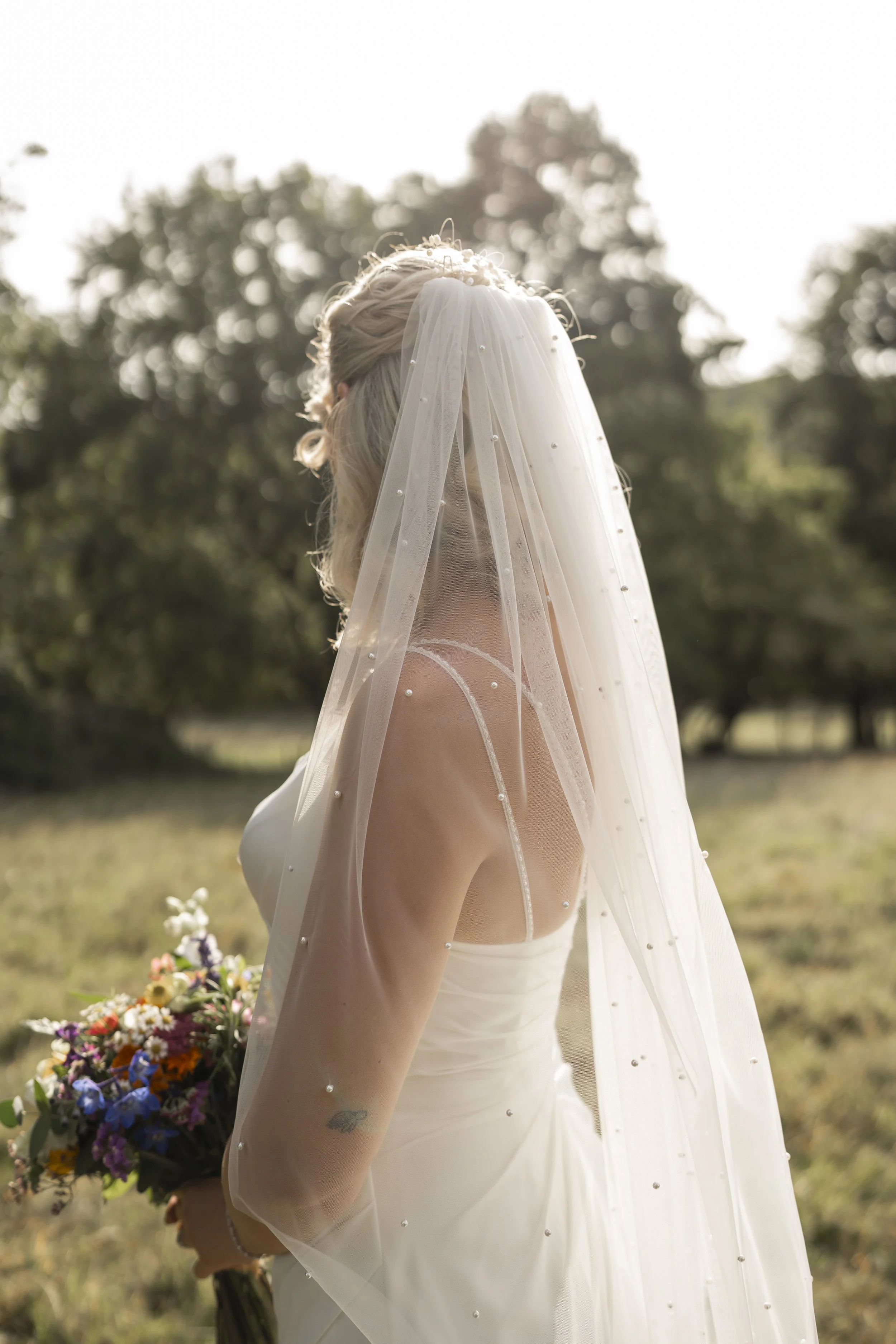 A bride with blonde hair wearing a white wedding gown and veil, holding a colorful bouquet, standing outdoors with trees in the background.