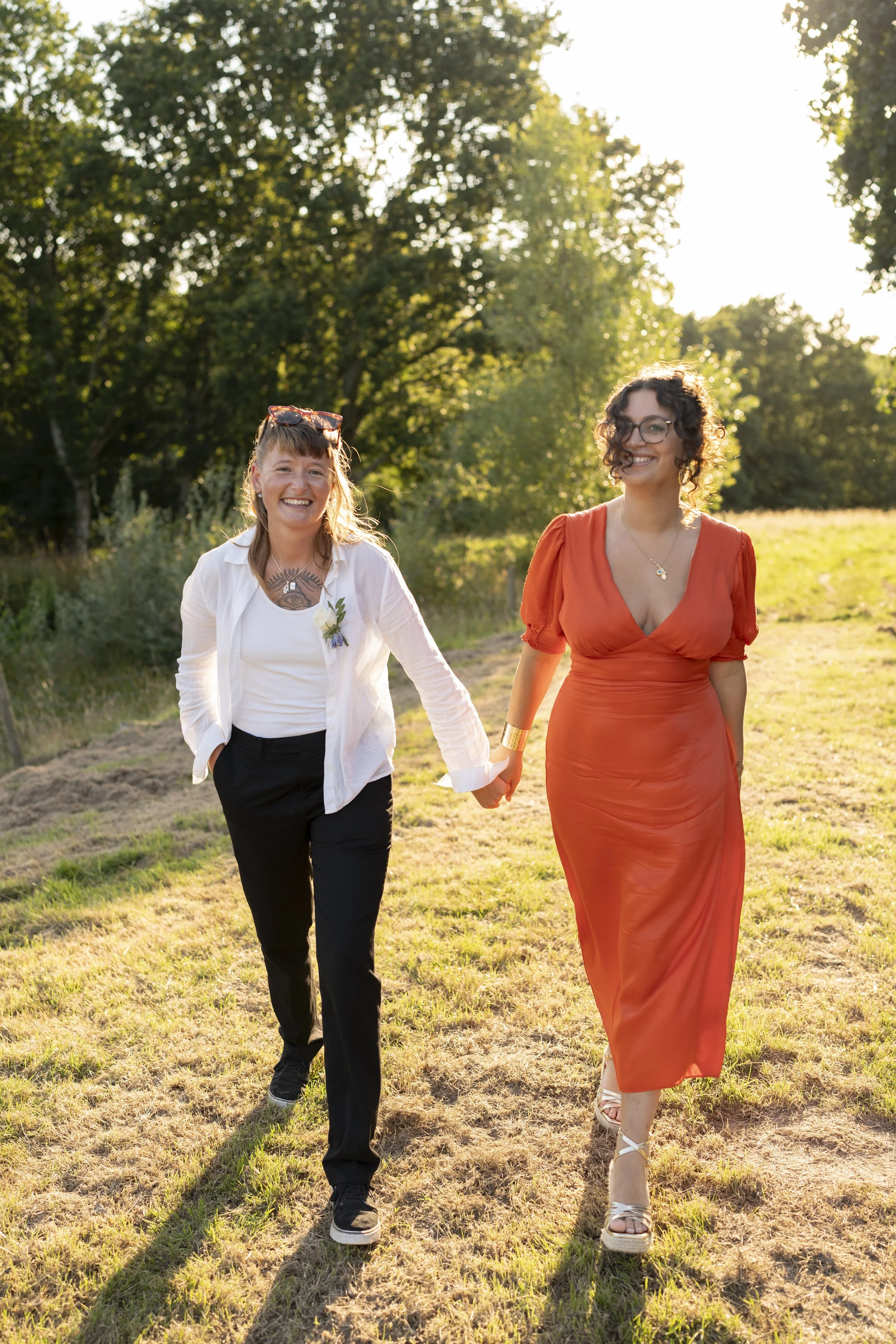 Two women walking hand in hand outdoors in a grassy field during sunset, smiling and enjoying each other's company.