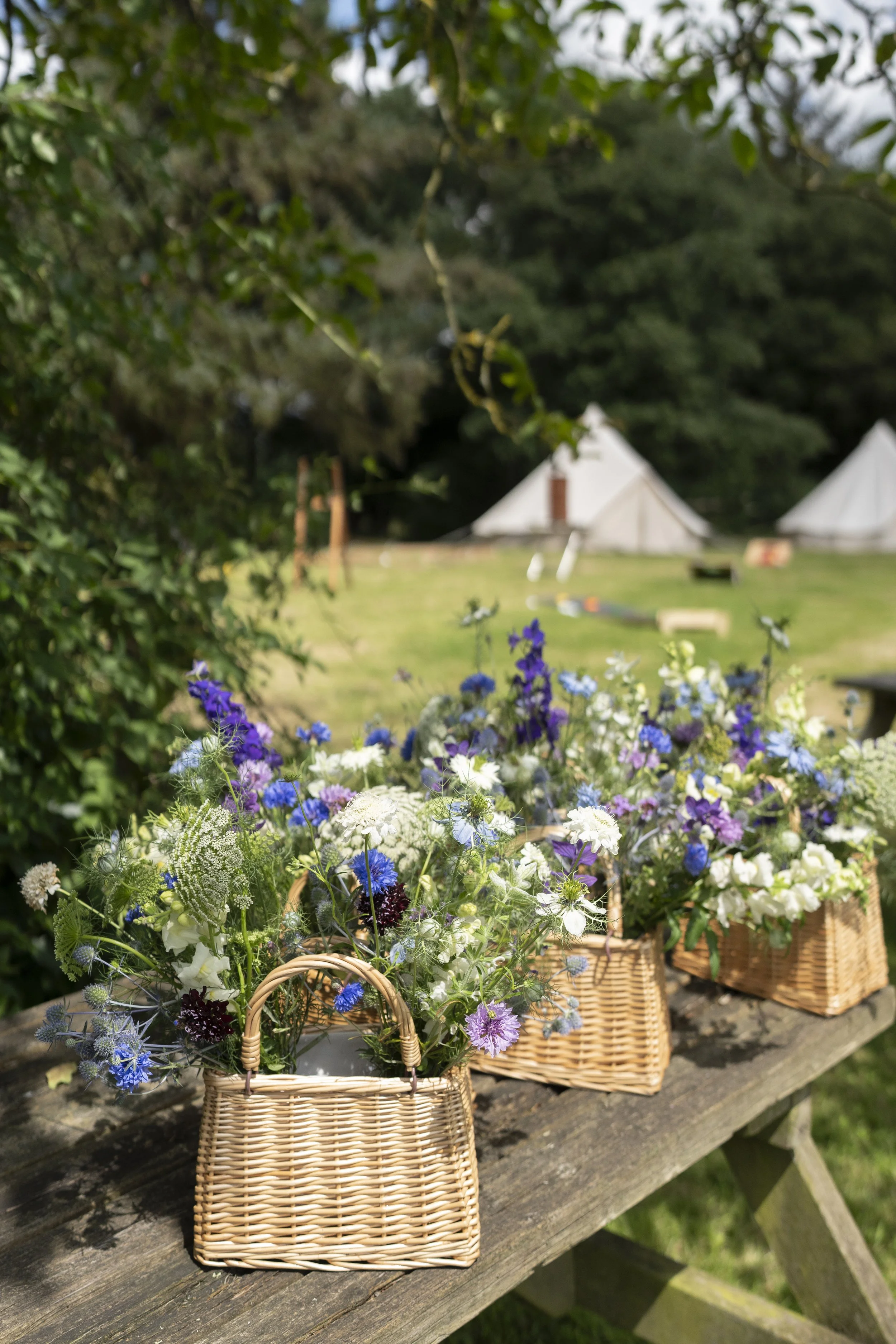 Multiple baskets filled with colorful flowers on a wooden table outdoors with tents and greenery in the background