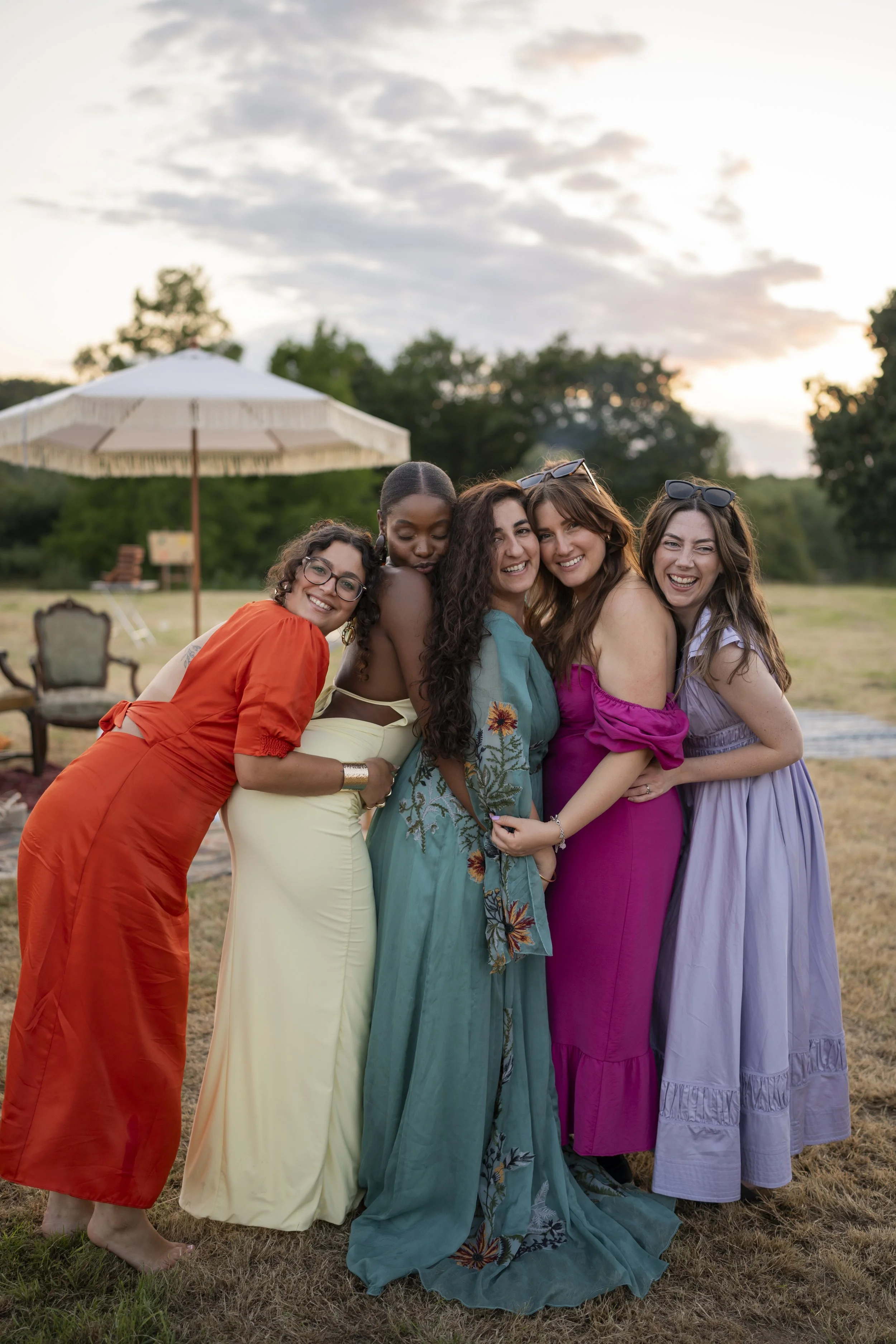 Five women in colorful dresses smiling and hugging outdoors at sunset with trees and a large umbrella in the background.