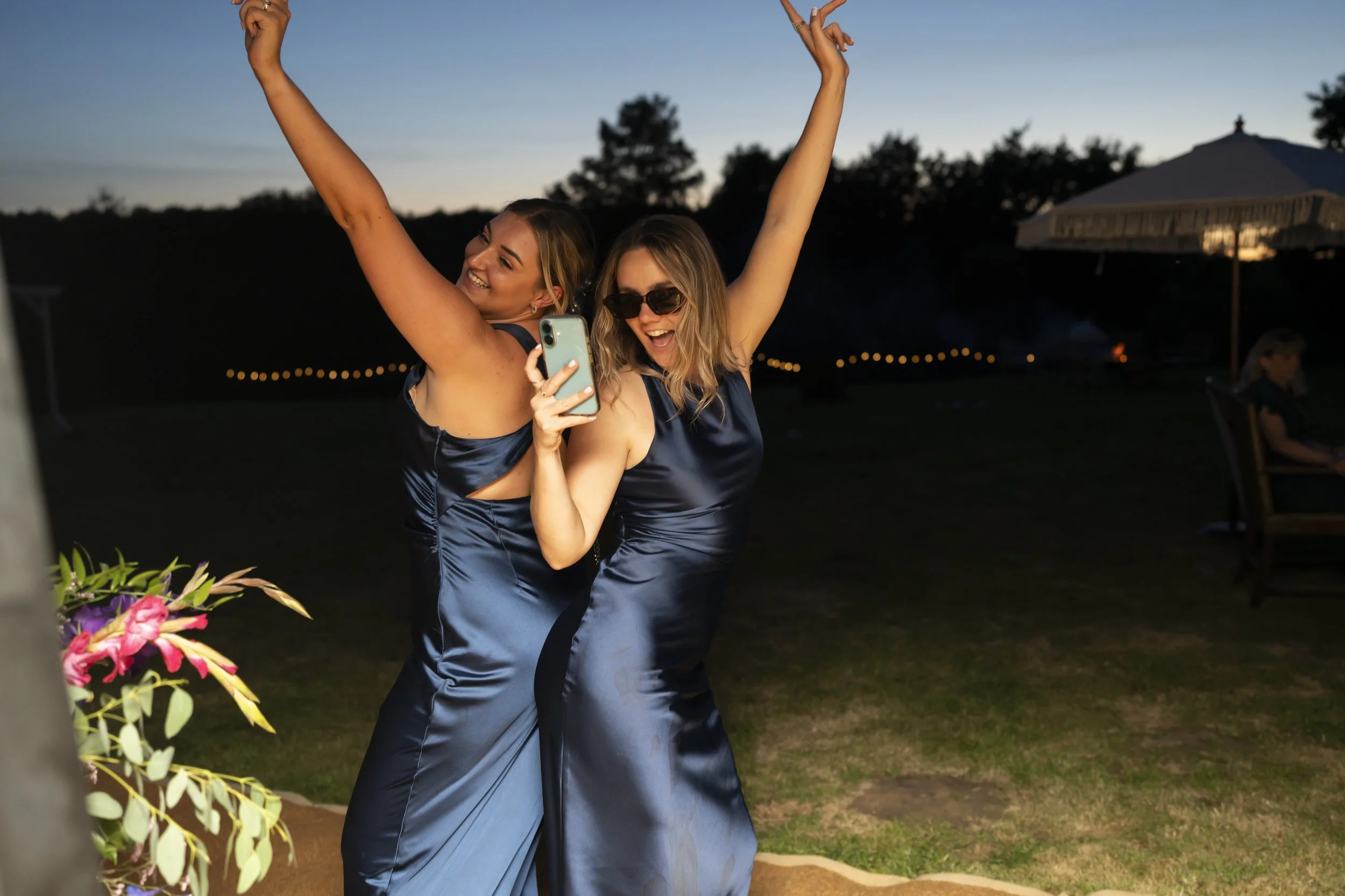 Two women in matching navy satin dresses celebrating outdoors at dusk, smiling, one taking a selfie, with trees, a tent, and string lights in the background.