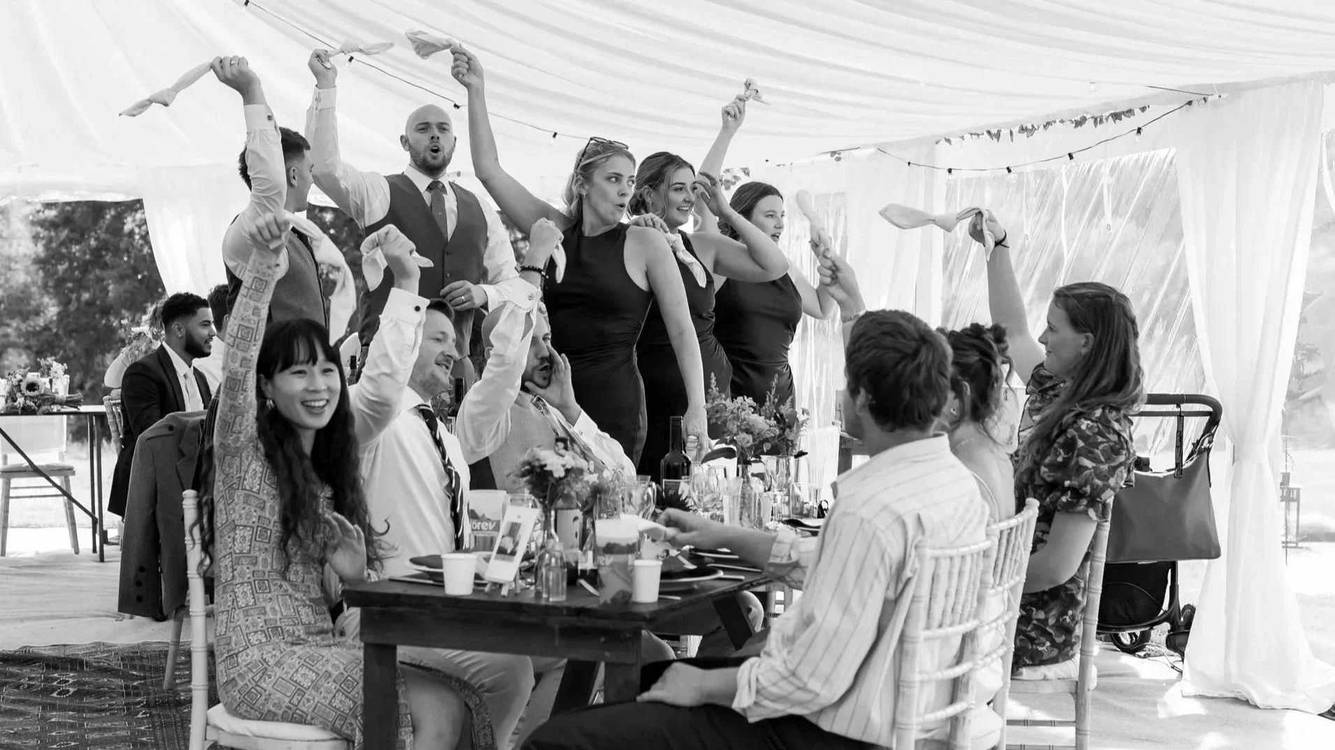 A group of people at a celebration or wedding under a tent, some standing and others sitting, with cheerful and celebratory expressions.