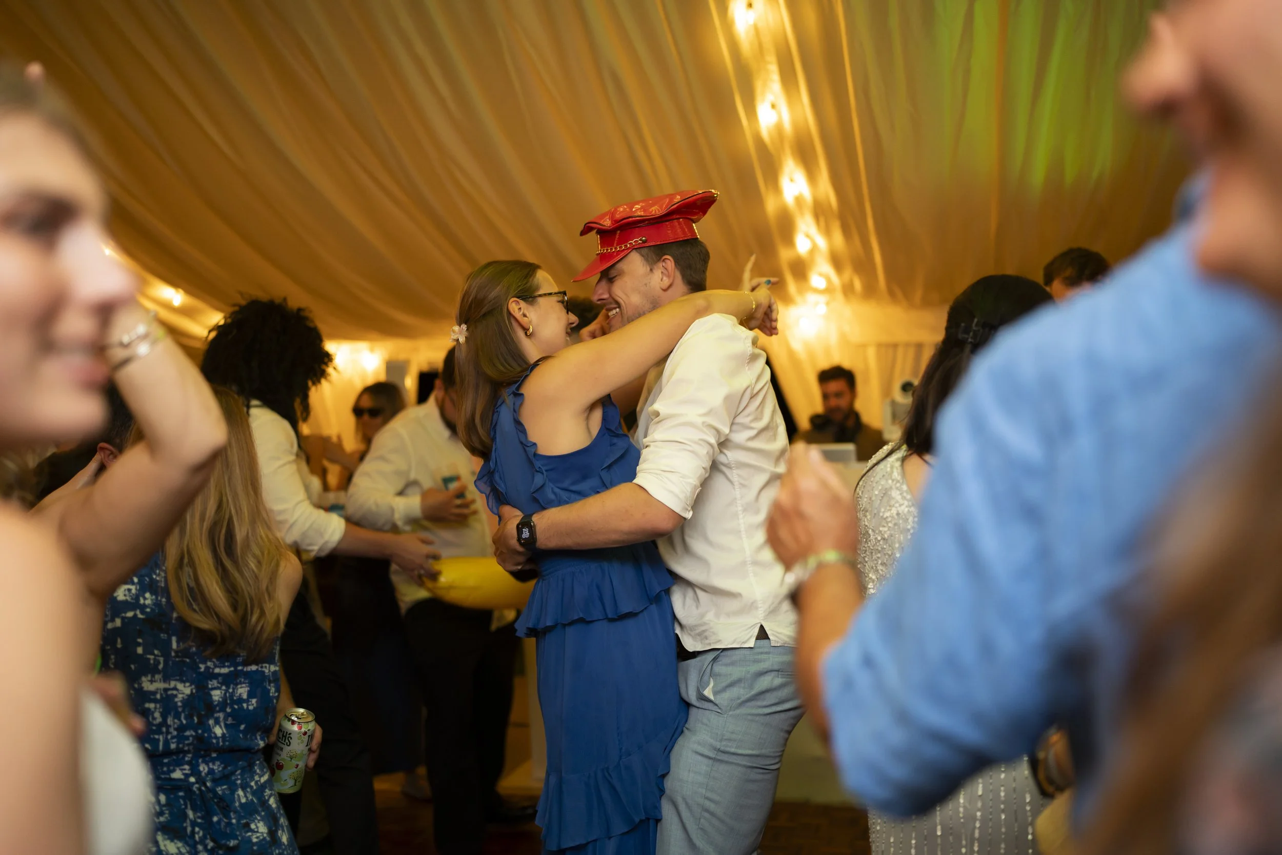 A couple dancing close together at a party or celebration, smiling and enjoying each other's company. The woman is wearing a blue dress, and the man is wearing a white shirt and has a red hat on his head. The background shows a warmly lit, decorated ceiling with other people in the distance.