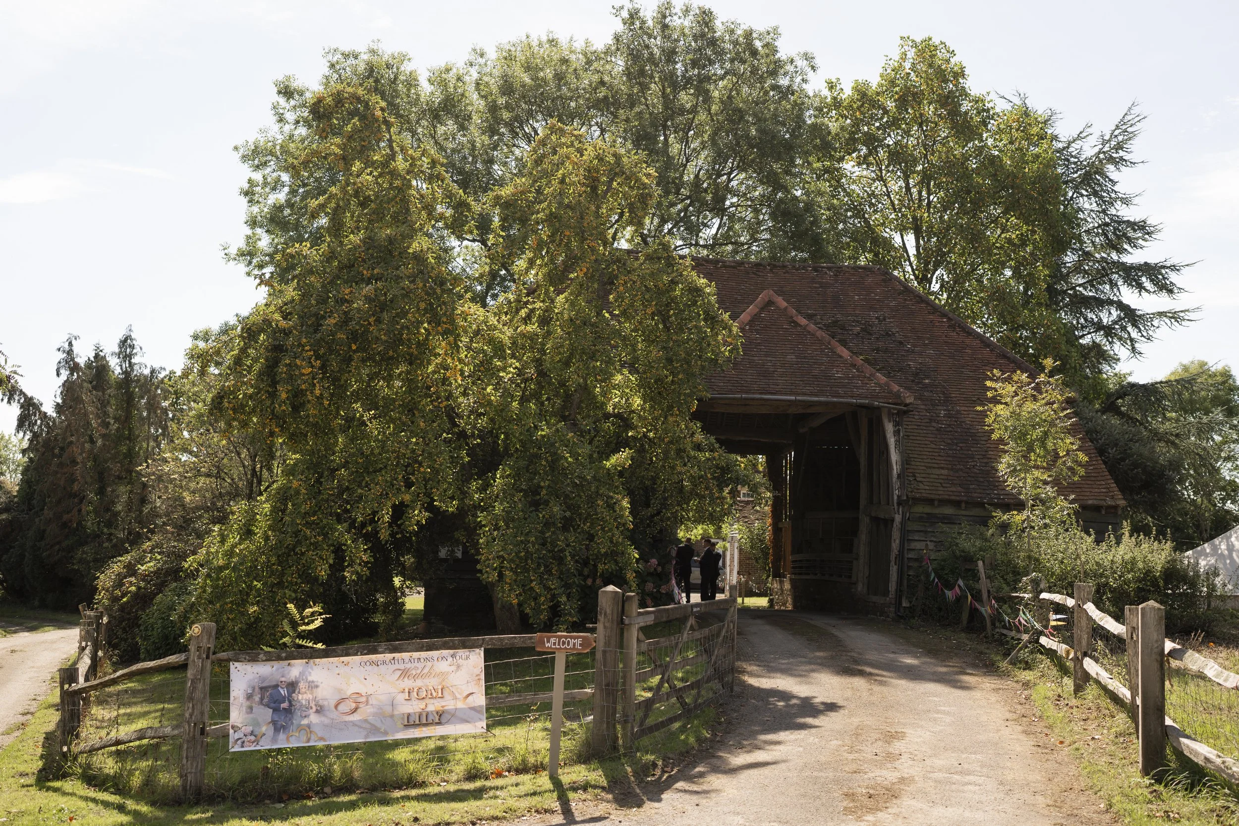 A rustic outdoor setting with an old wooden barn, large trees, and a sign announcing a wedding celebration for Tom and Lily.