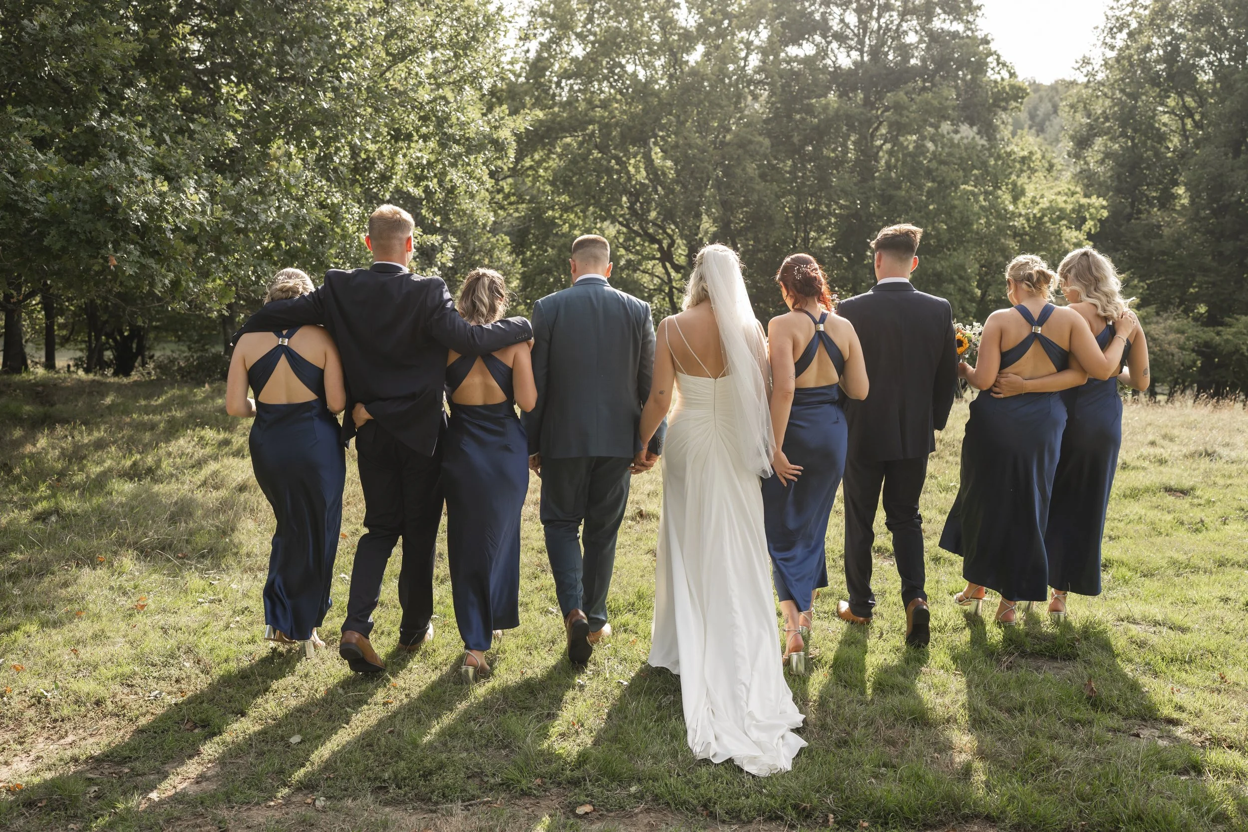 Group of wedding guests, including bridesmaids and groomsmen, walking outdoors hand-in-hand, with a bride in a white gown at the center, surrounded by green trees.