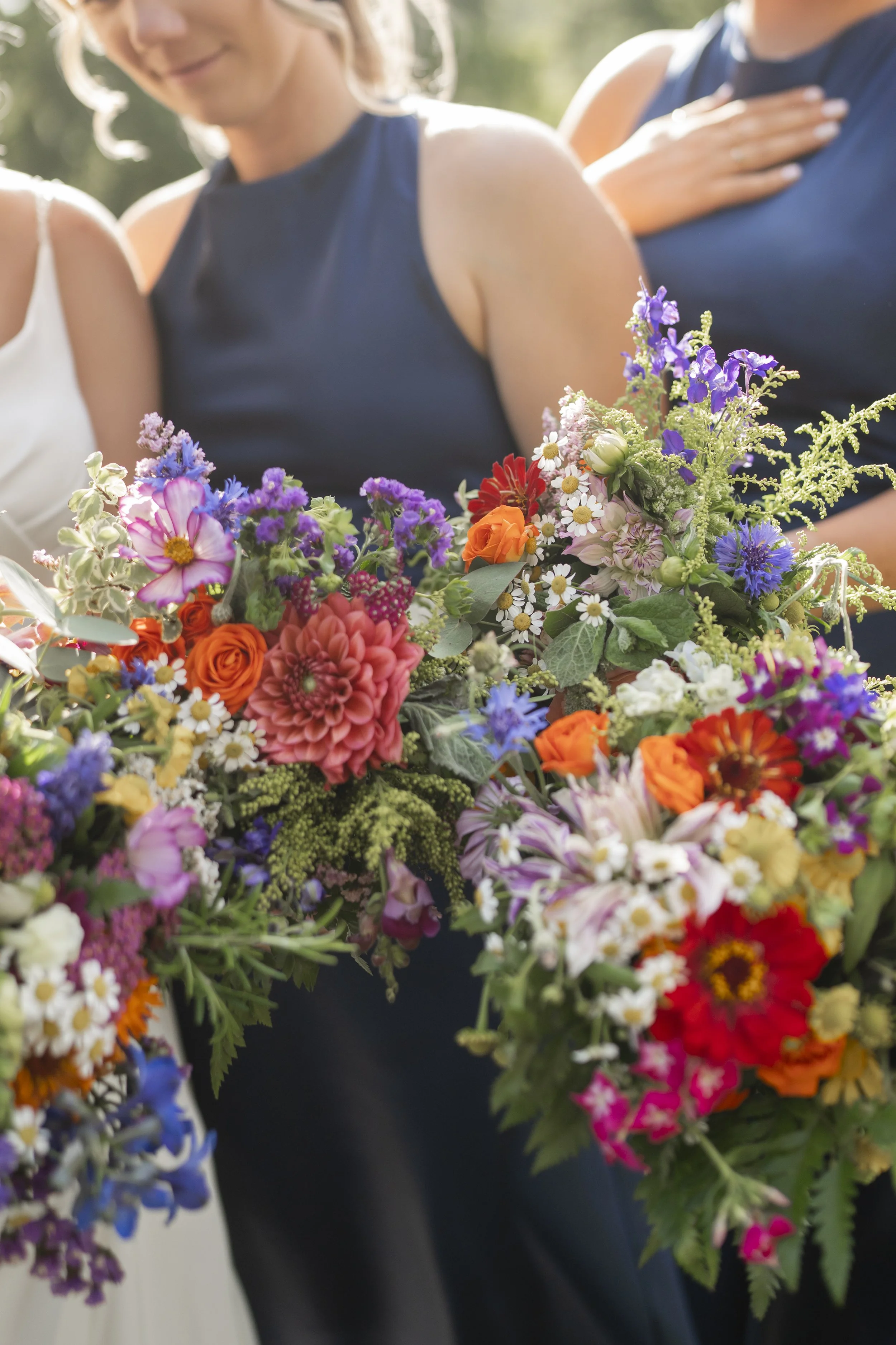 A person wearing a dark sleeveless dress stands outdoors with other individuals, holding a large bouquet of multicolored flowers including pink, purple, orange, and white blossoms.