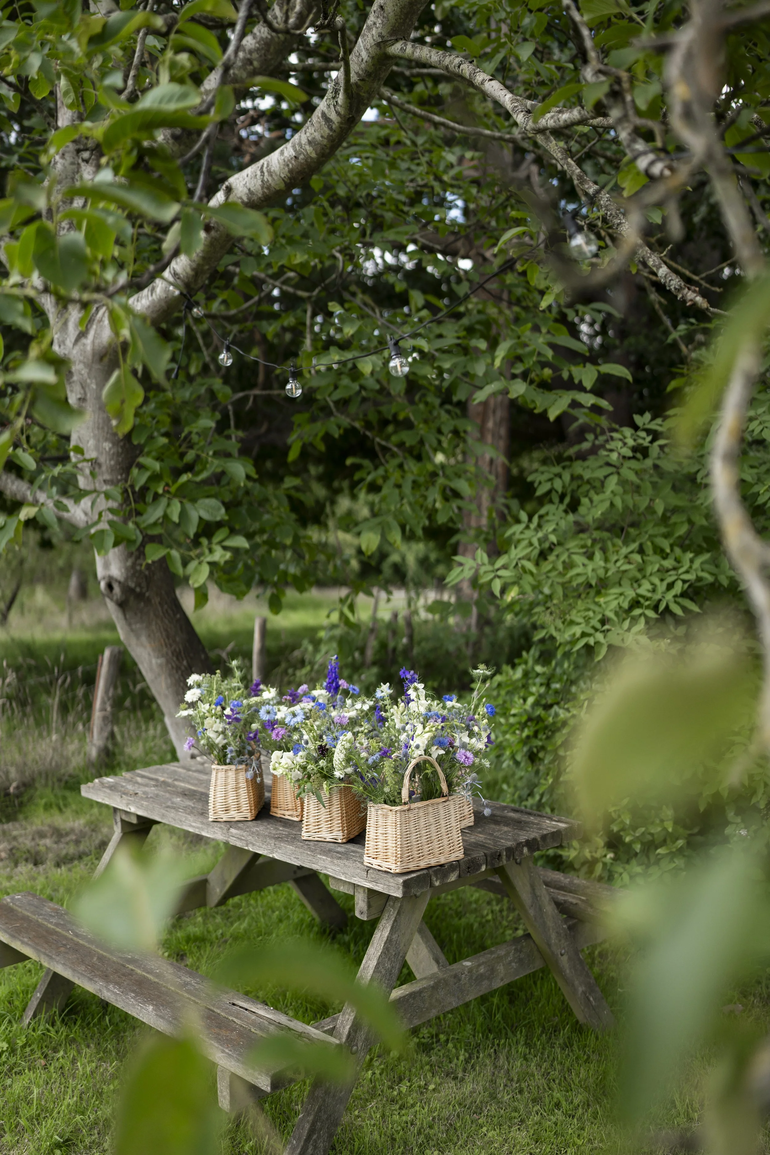 A rustic wooden picnic table outdoors under a tree, decorated with woven baskets of colorful flowers, with string lights hanging among the branches.