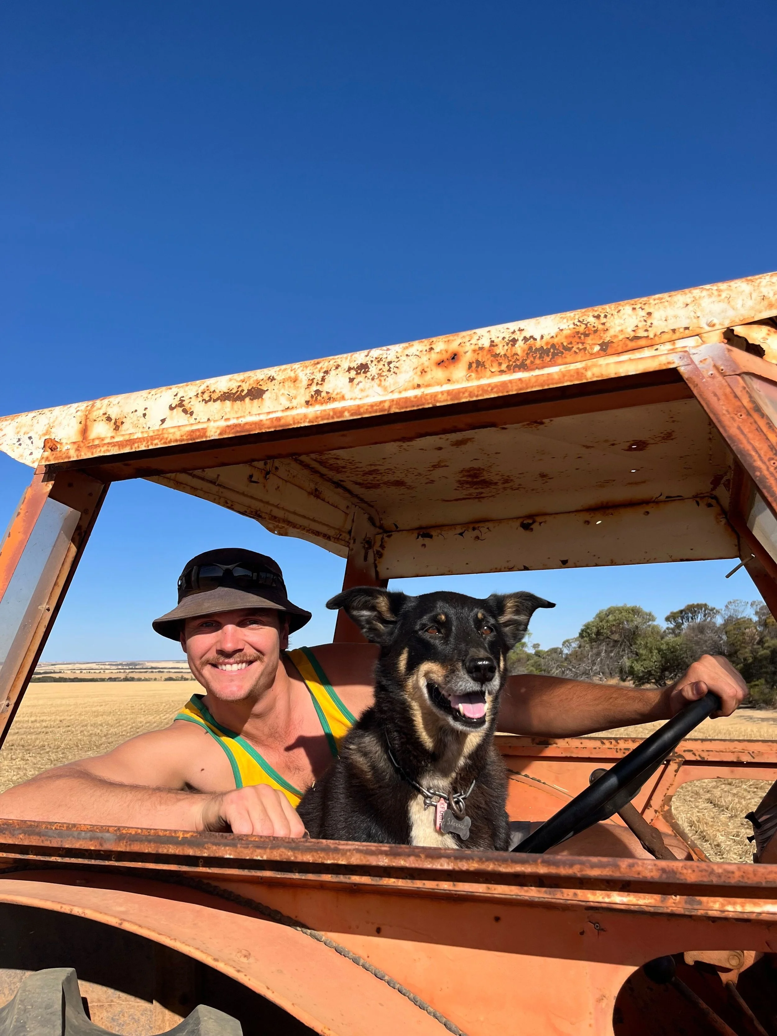 A man with a wide-brimmed hat and sleeveless shirt smiling and leaning out of an old, rusty tractor with a black and tan dog sitting inside. The background shows an open field and a blue sky.