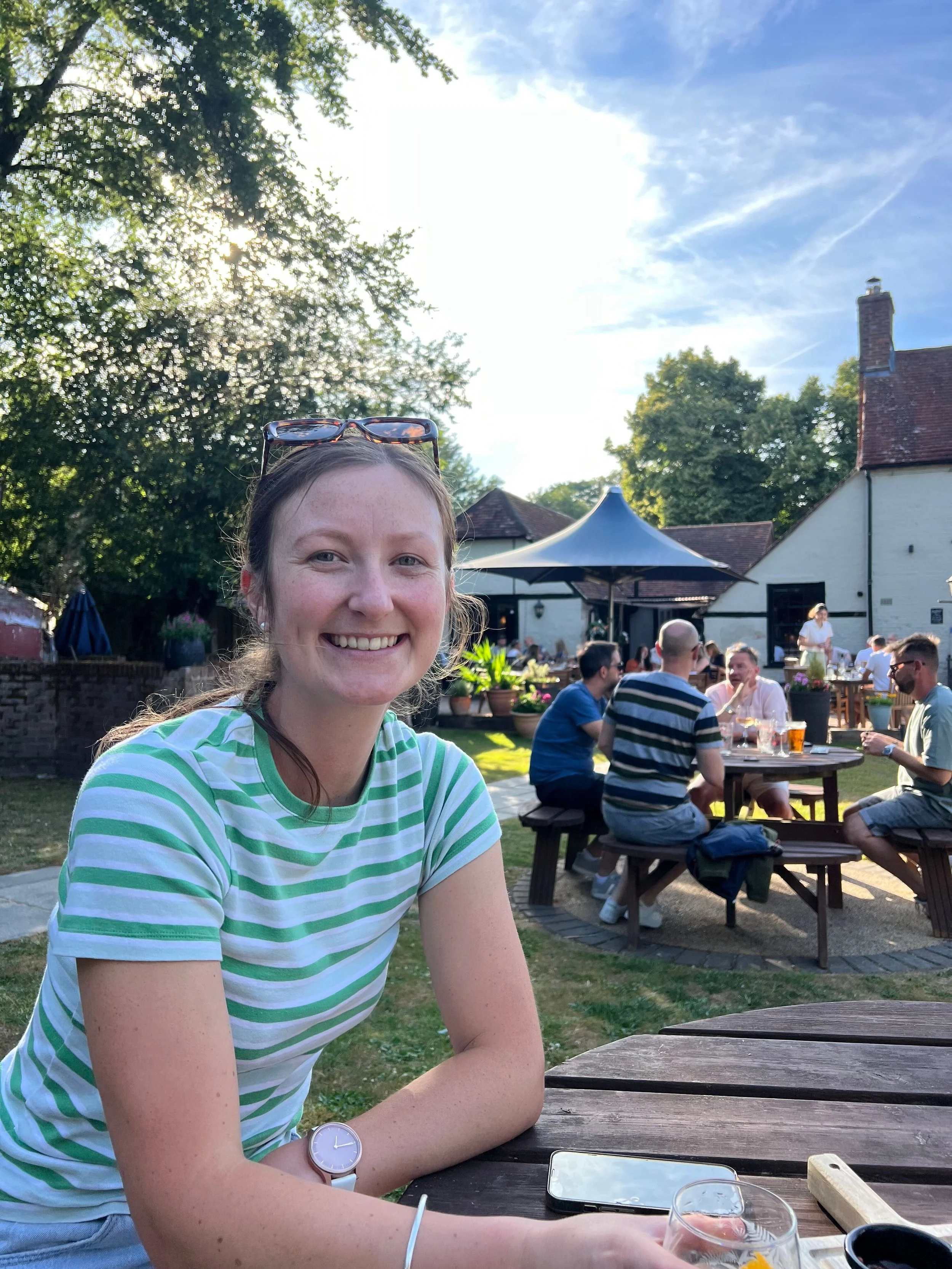 A smiling woman with sunglasses resting on her head, wearing a green and white striped t-shirt, sitting outdoors at a wooden table during daytime. In the background, there are people socializing, potted plants, a large umbrella, trees, and a building with a chimney under a blue sky with some clouds.