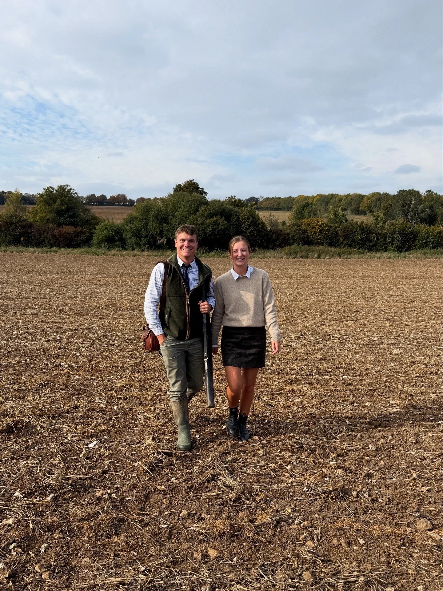 Two smiling young adults walking in a plowed field with trees and a cloudy sky in the background.