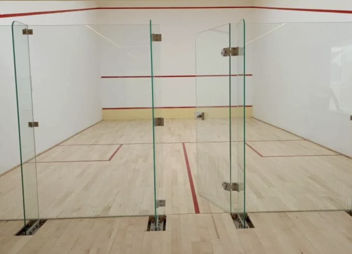 Indoor squash court with glass walls, light wood flooring, and red court markings