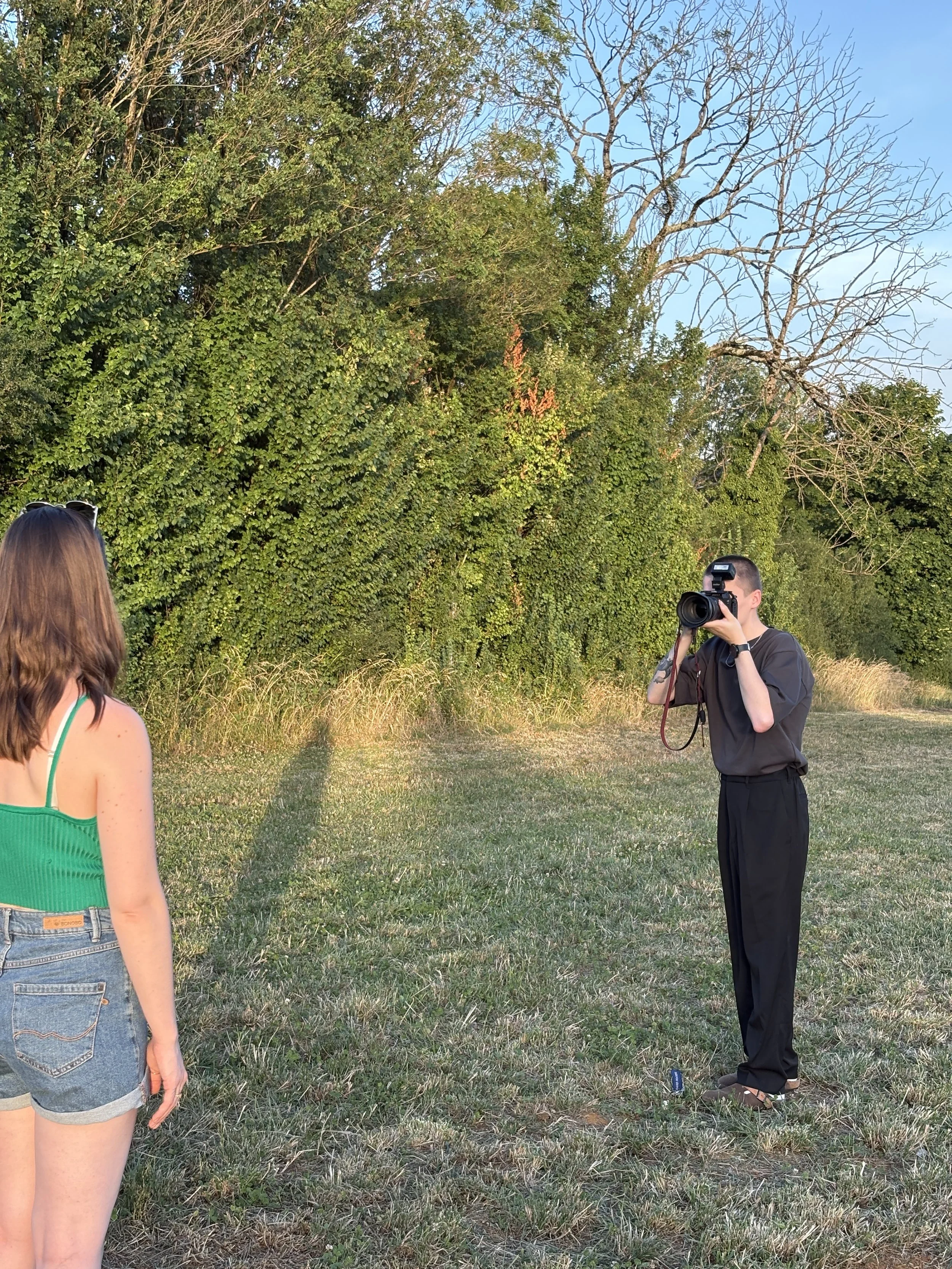 Une femme avec un appareil photo regarde un groupe de personnes dans une zone herbeuse avec des arbres, certains arbres étant sans feuilles, sous un ciel clair.