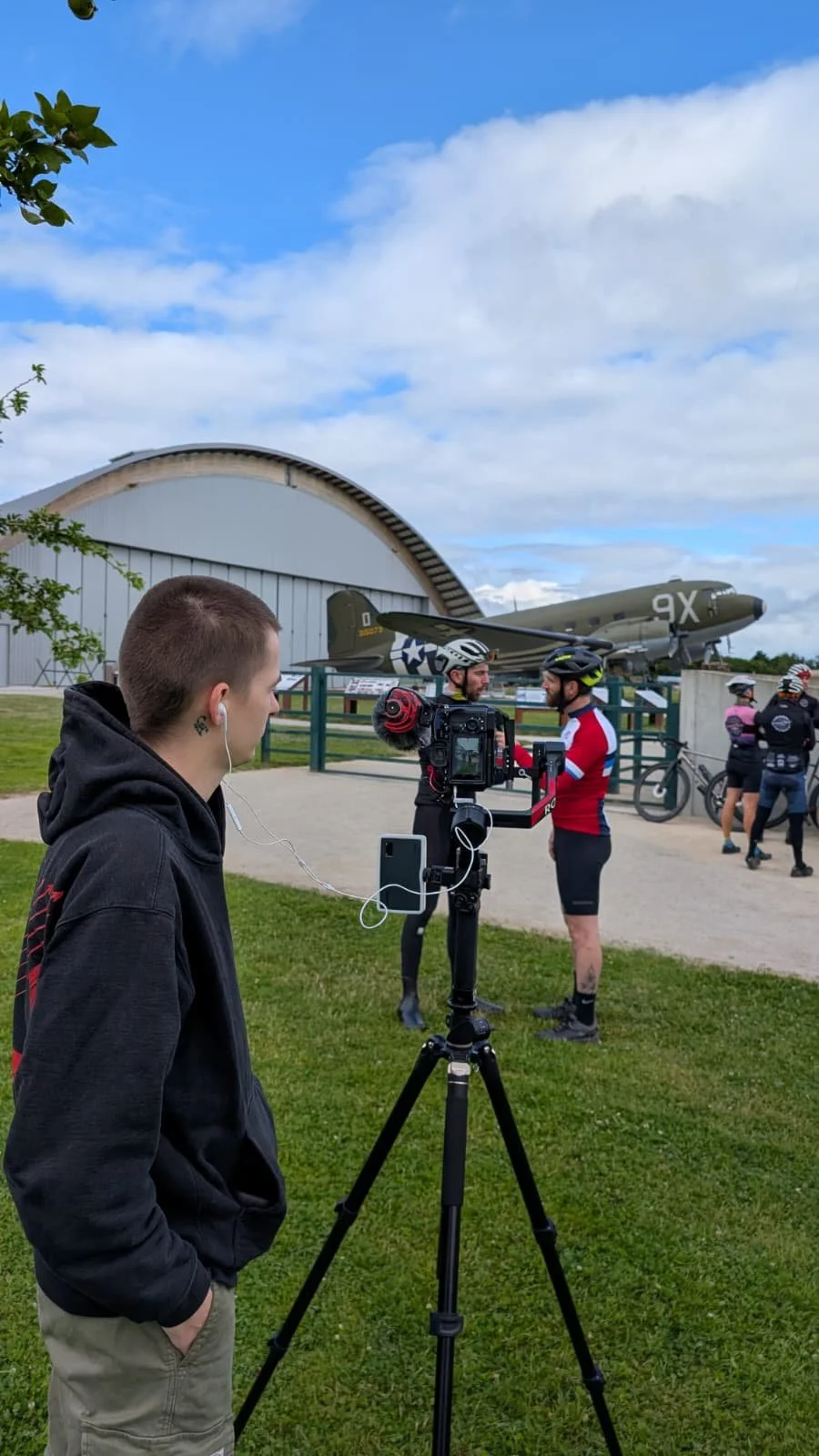 Un jeune homme observe un tournage avec une caméra sur un trépied, plusieurs cyclistes s'entraînent ou discutent devant un avion de la Seconde Guerre mondiale exposé à l'extérieur, sous un ciel partiellement nuageux.