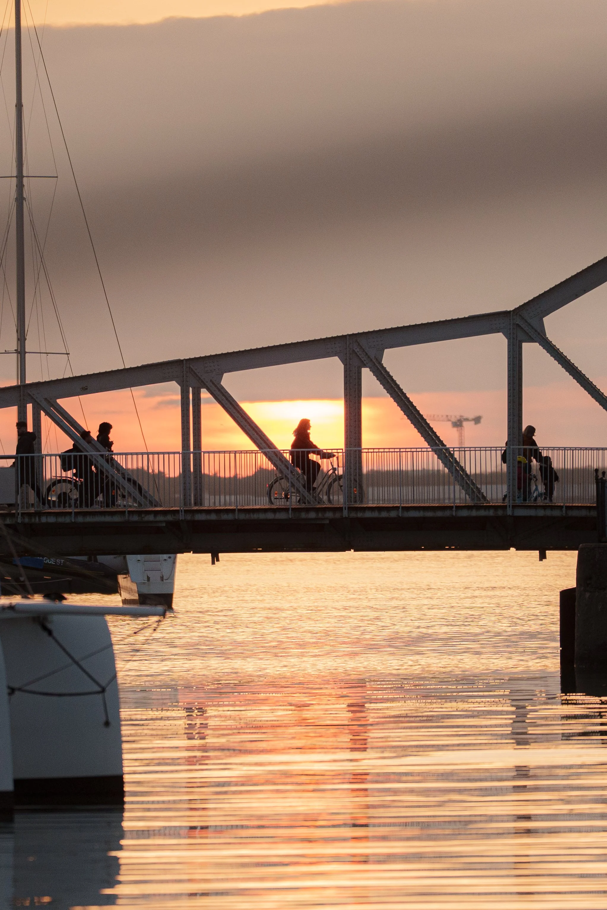 Coucher de soleil sur un pont où circulent des personnes à vélo et à pied, reflection dans l'eau, avec des navires au quai.