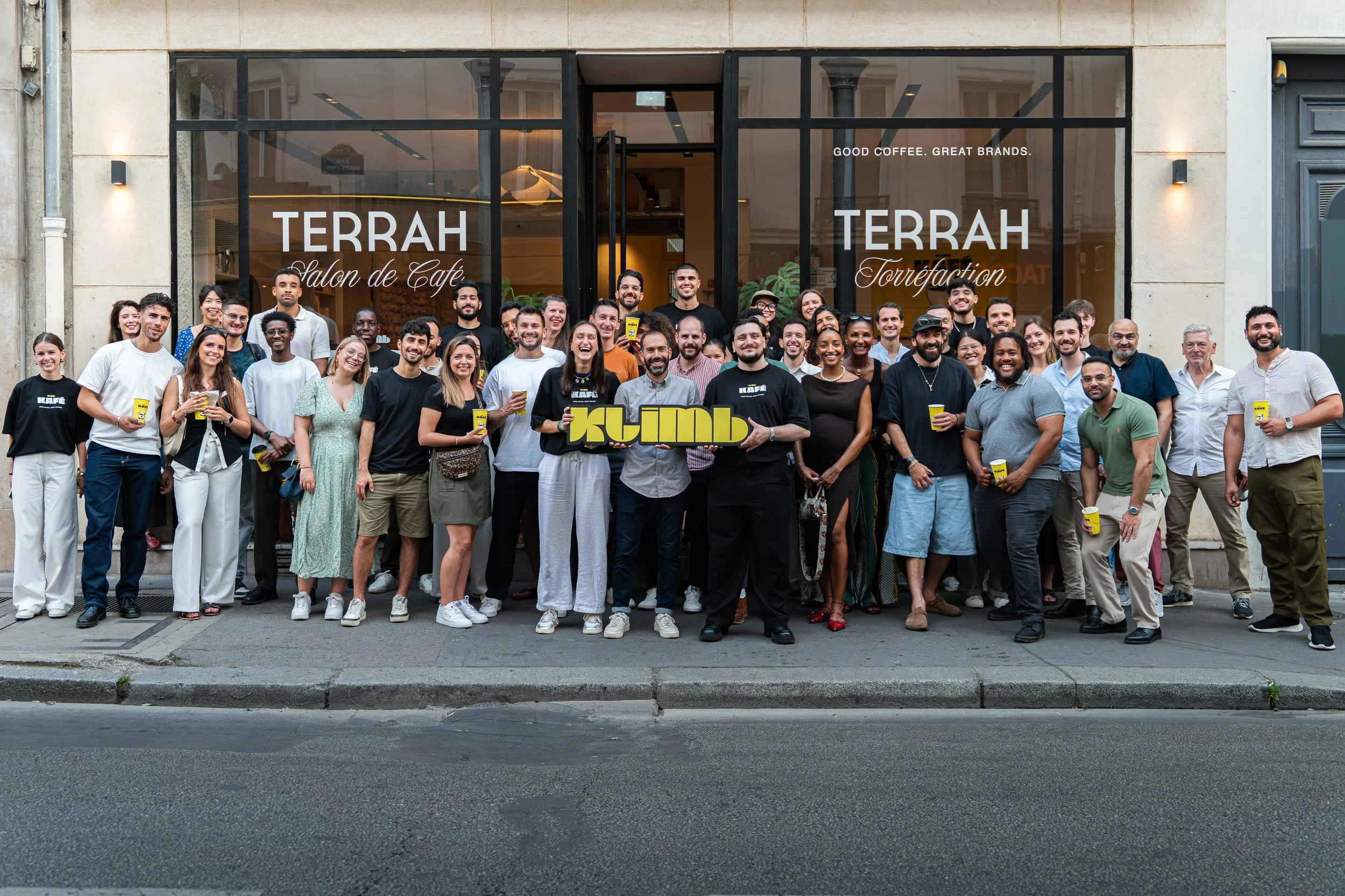 Un groupe diversifié de personnes posant devant un café nommé 'TERRAH Salon de Café', certains tenant des gobelets de café, lors d'une sortie ou événement communautaire.
