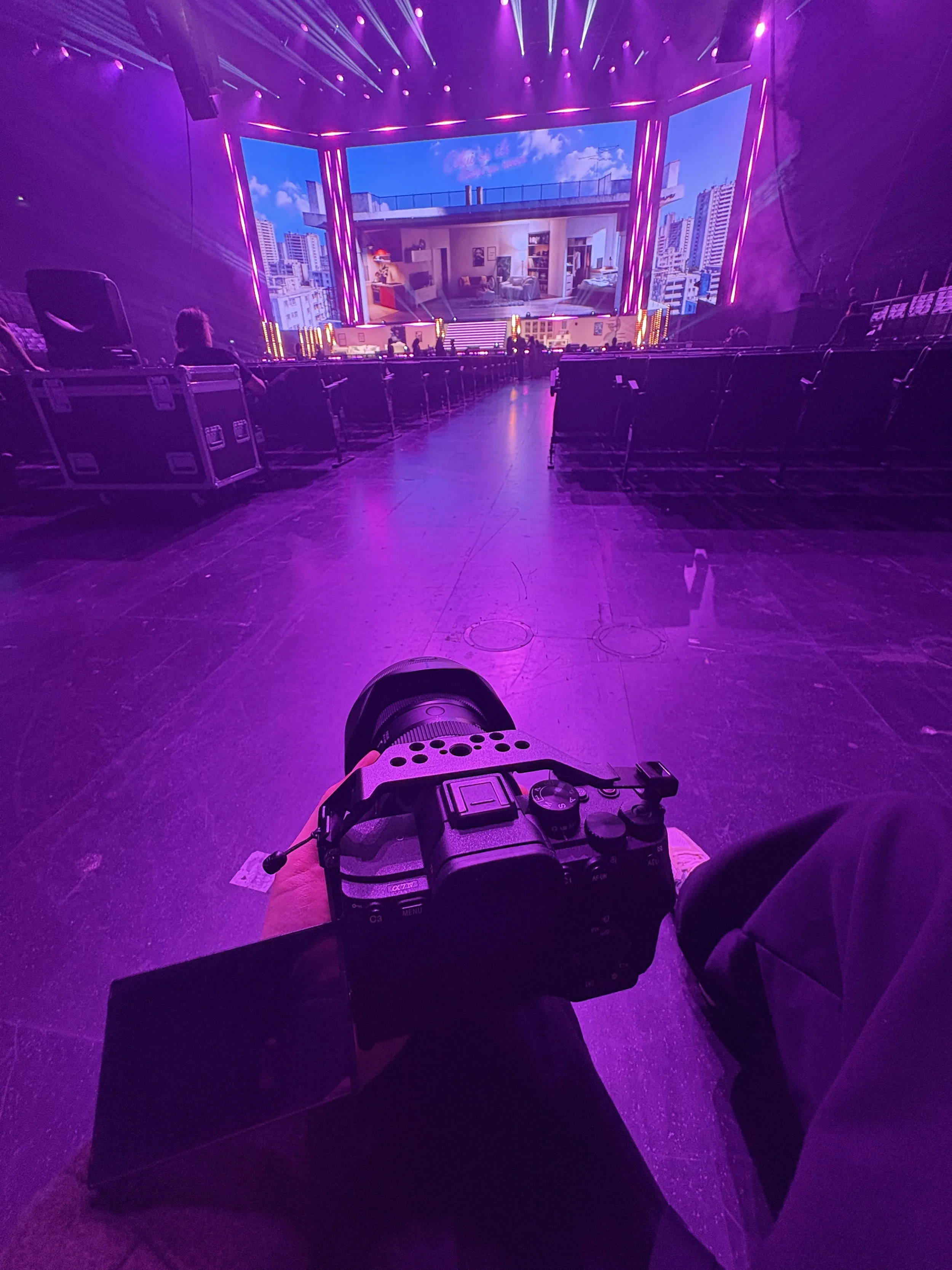 Photo prise d'une salle de spectacle ou auditorium avec des sièges vides, un écran géant affichant une image d'un appartement, et un éclairage violet. Au premier plan, une personne tient une caméra numérique.