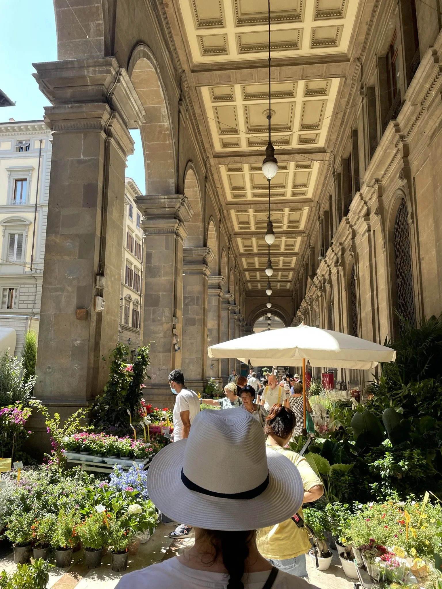 A woman wearing a large white sunhat with a black band, viewed from behind, shopping at an outdoor flower market with multiple people and vibrant flower displays under a covered walkway with ornate architecture.