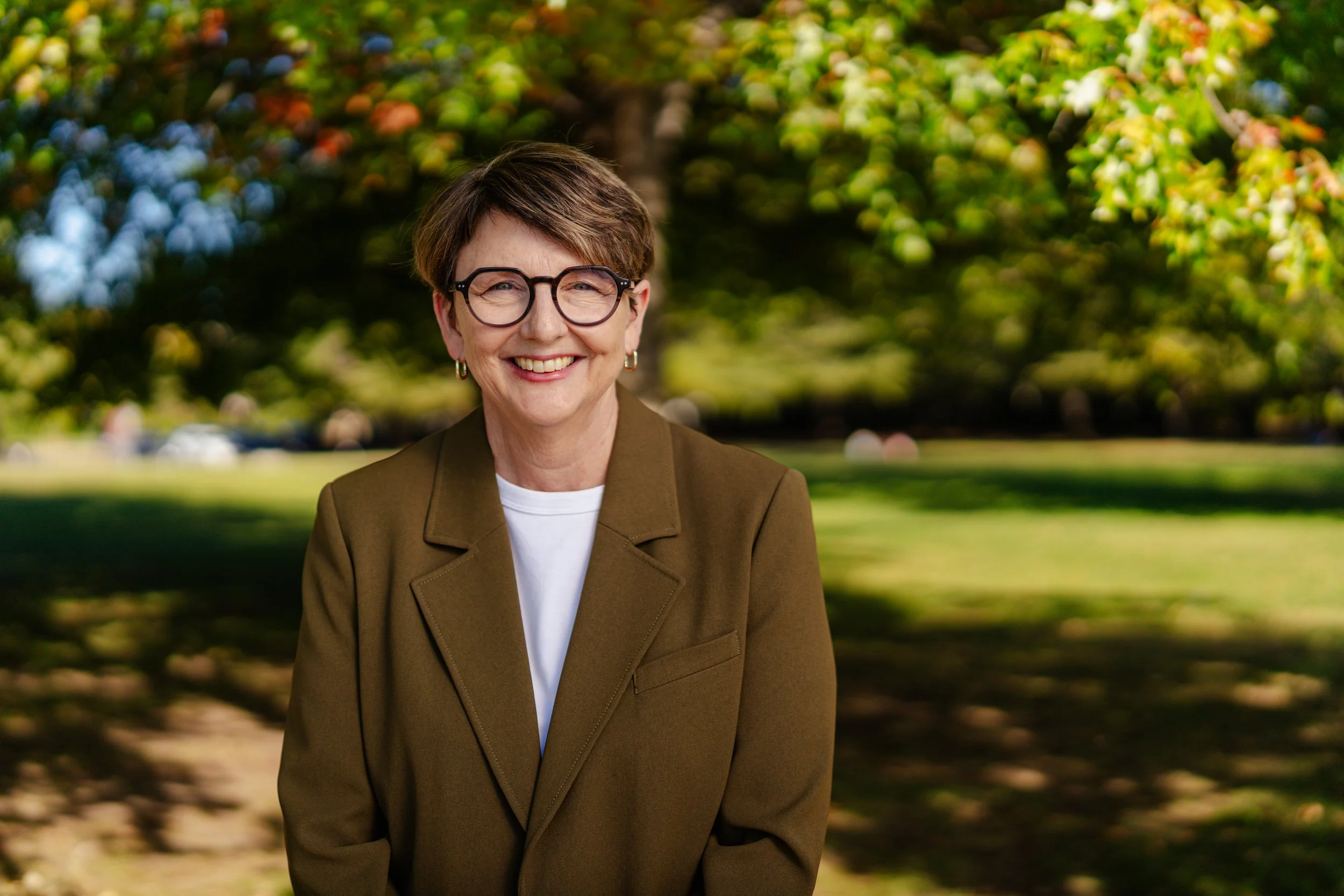 A middle-aged woman with short brown hair, wearing glasses and earrings, smiling outdoors in front of a park with green trees.