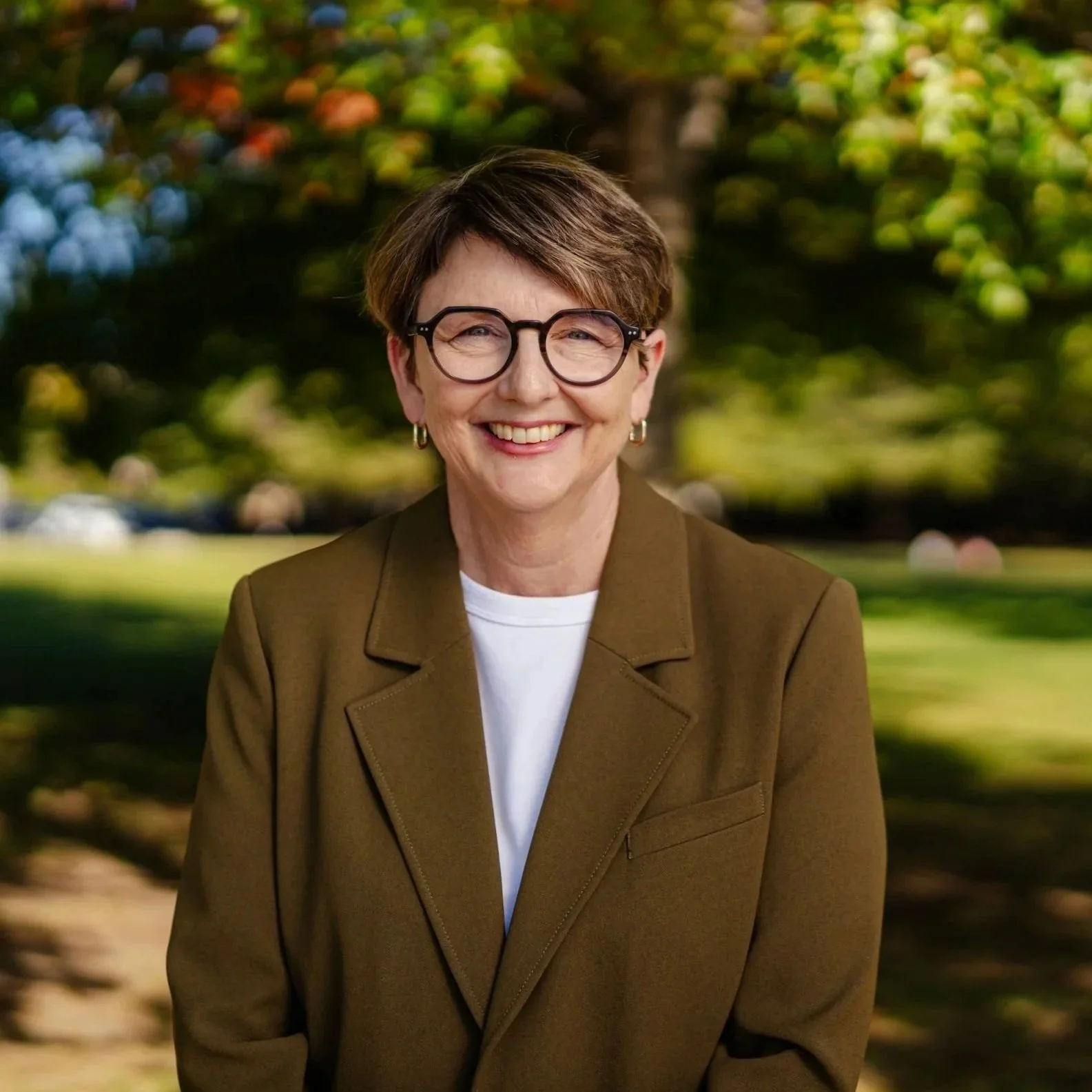 Smiling woman with short brown hair and glasses, wearing a brown blazer over a white shirt, outdoors with blurred green and yellow foliage in the background.