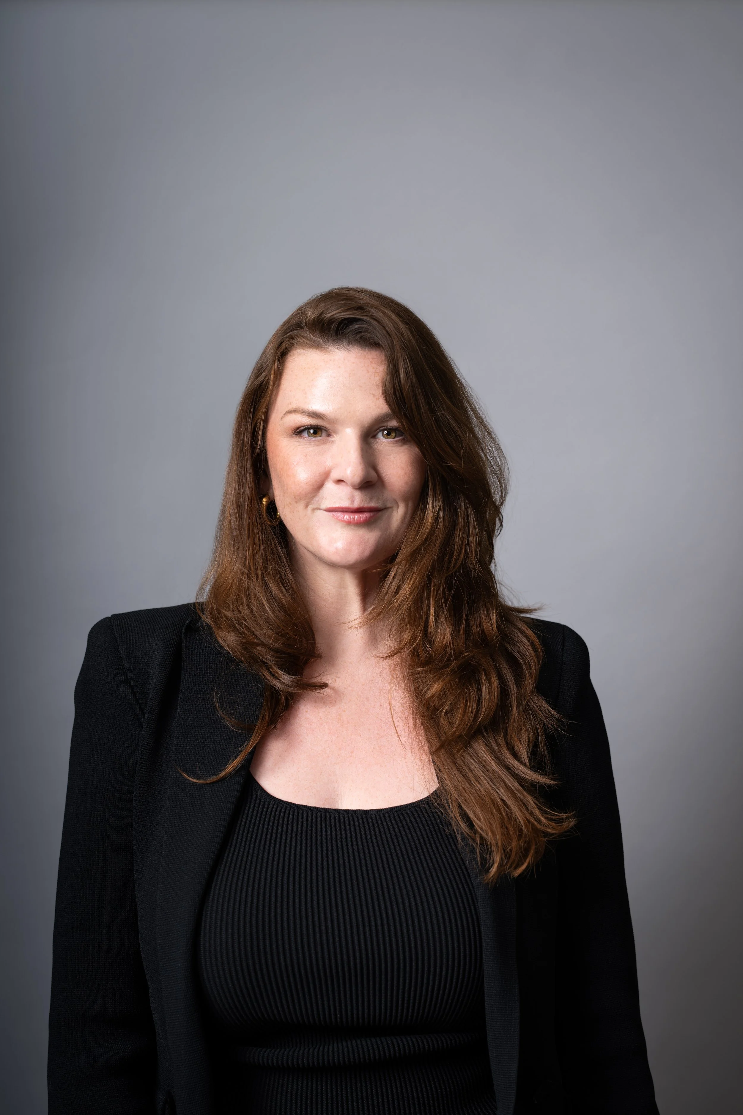 A woman with long brown hair, wearing a black blazer and black top, smiling slightly, against a gray background.
