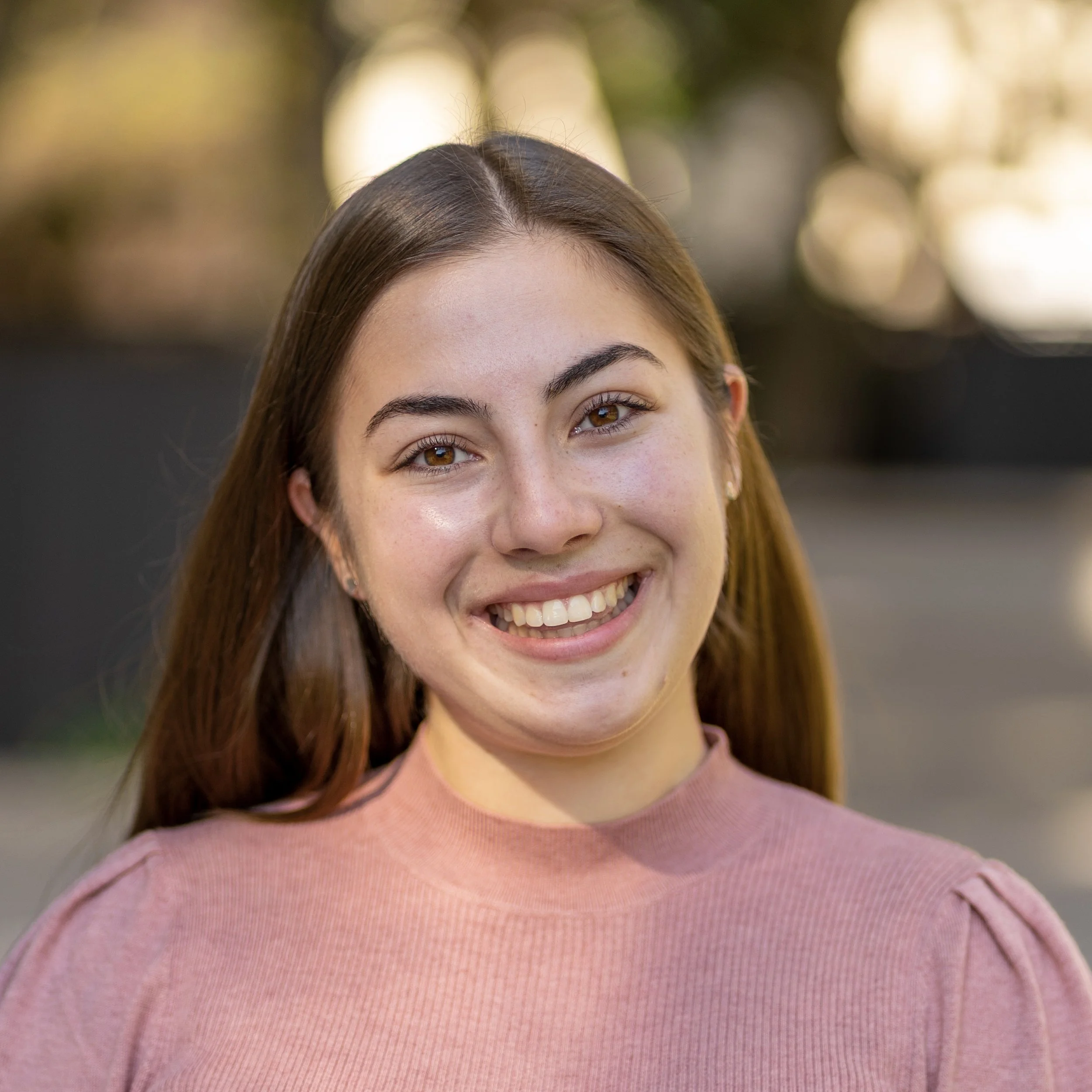 Close-up of a smiling woman with straight brown hair, wearing a pink sweater, outdoors with blurred background lights.