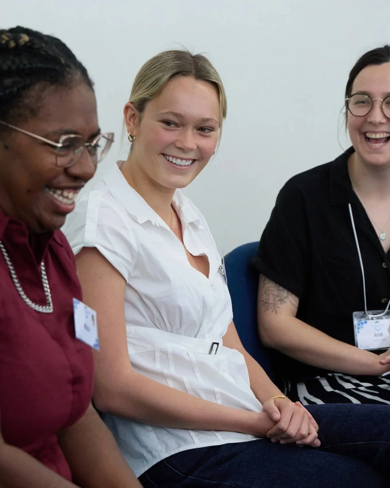 Three women sitting and smiling during a discussion or panel event, two of them wearing name tags, indoors with plain white wall background.