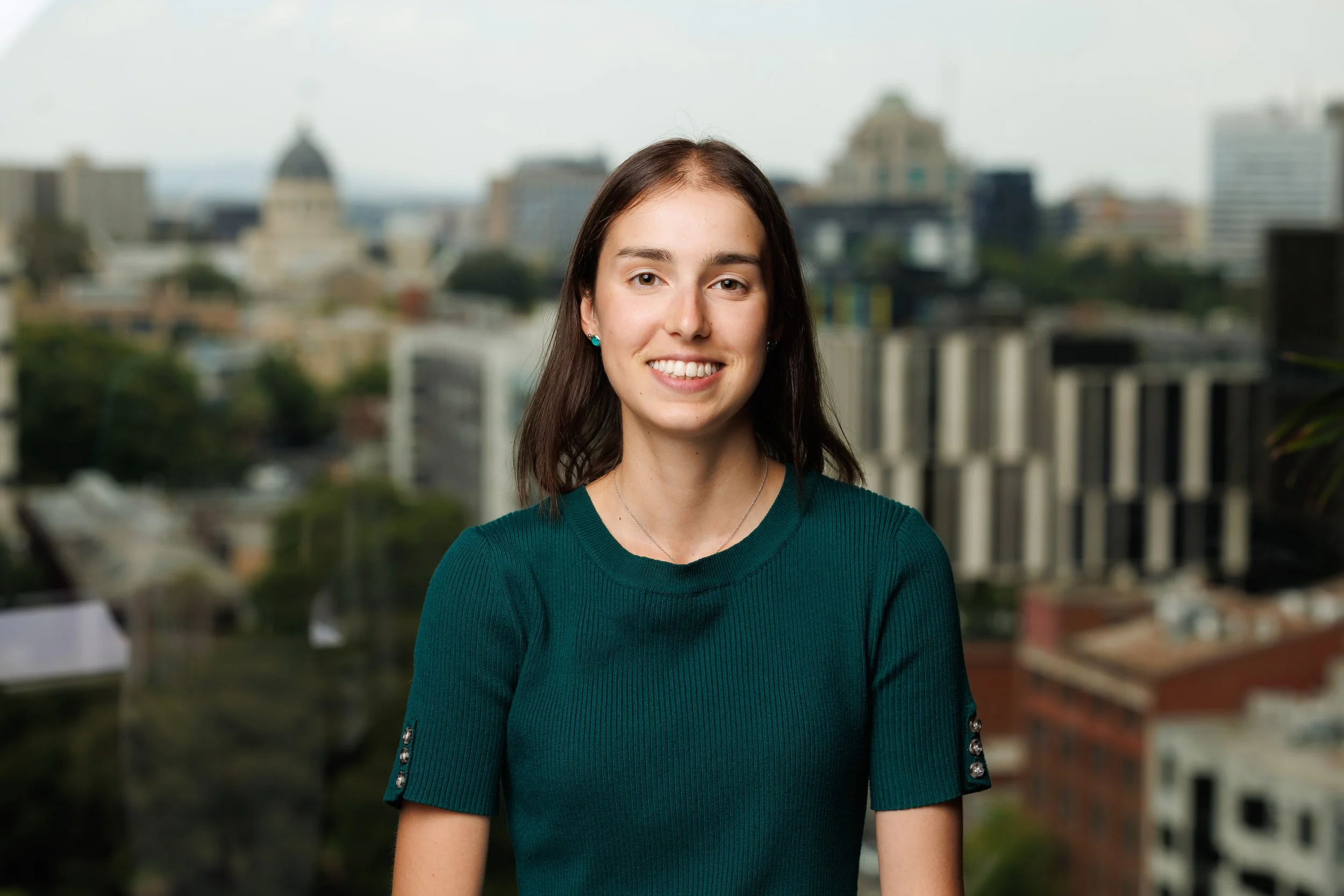A young woman with long dark hair, smiling, wearing a dark green sweater with decorative buttons on the sleeves, standing outdoors with a blurred cityscape in the background.