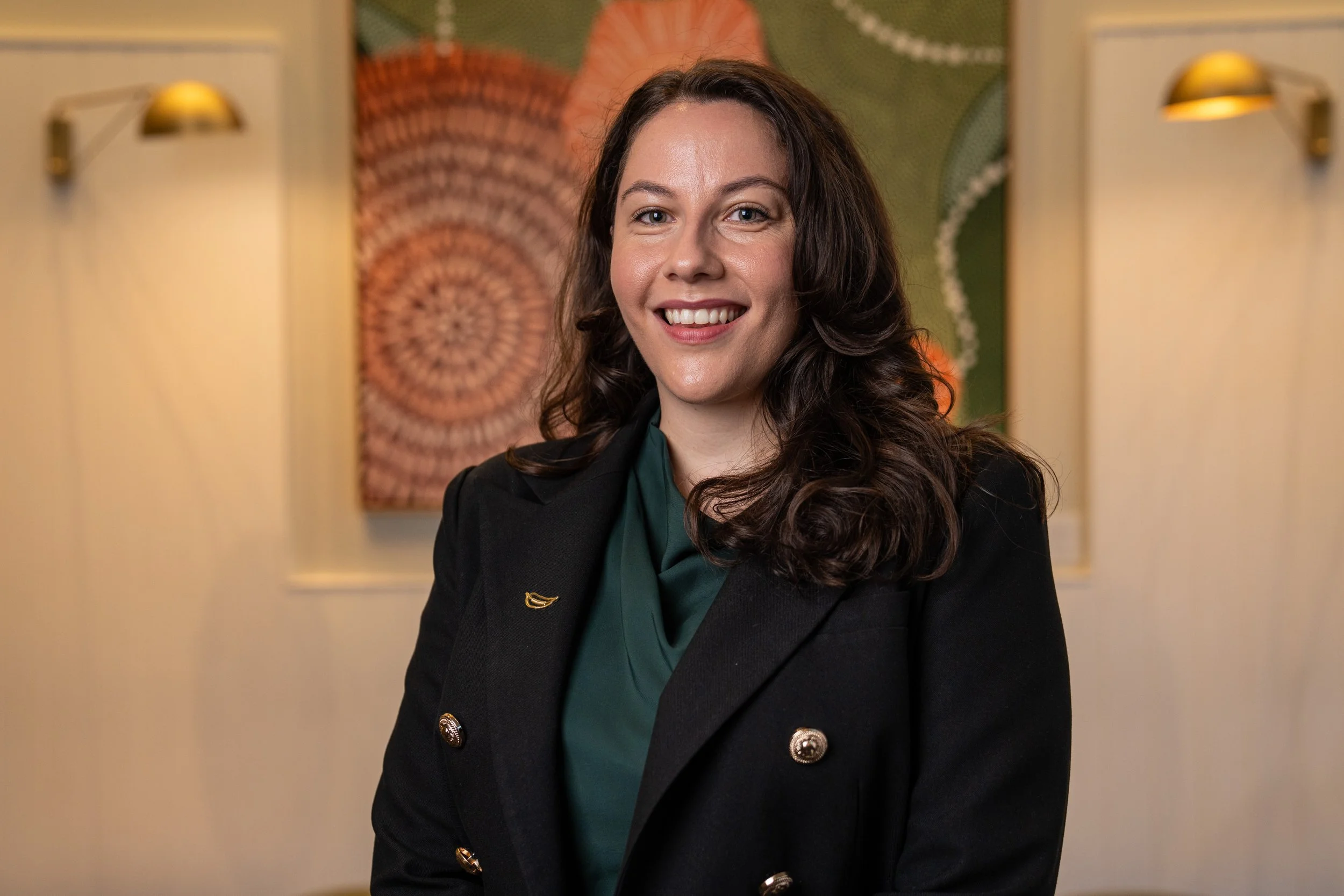 A smiling woman with long, dark, wavy hair wearing a black blazer with gold buttons and a green blouse, standing in front of colorful, abstract wall art.