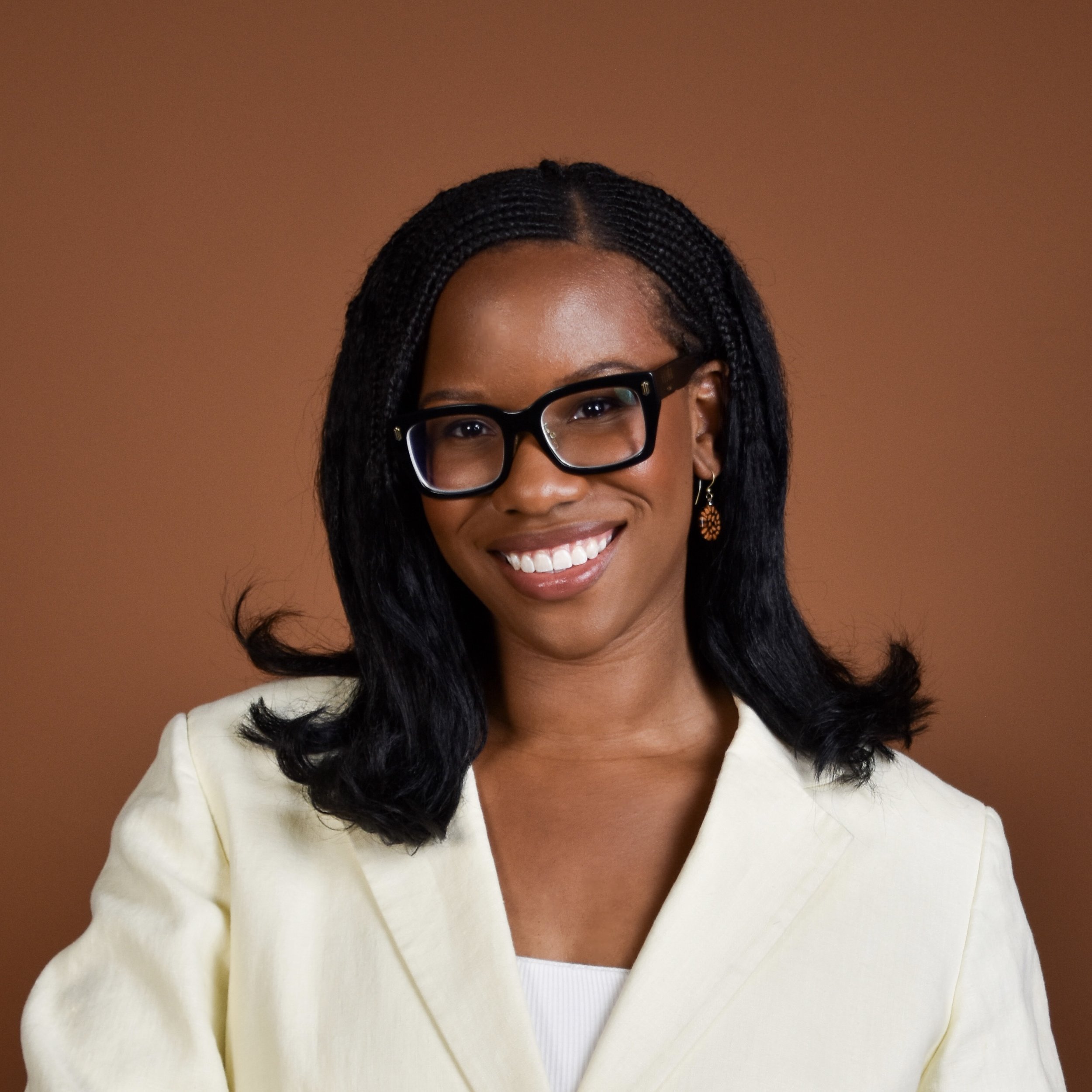 A woman with dark, braided hair, wearing glasses with black frames, earrings, a white top, and a cream-colored blazer, smiling against a plain brown background.