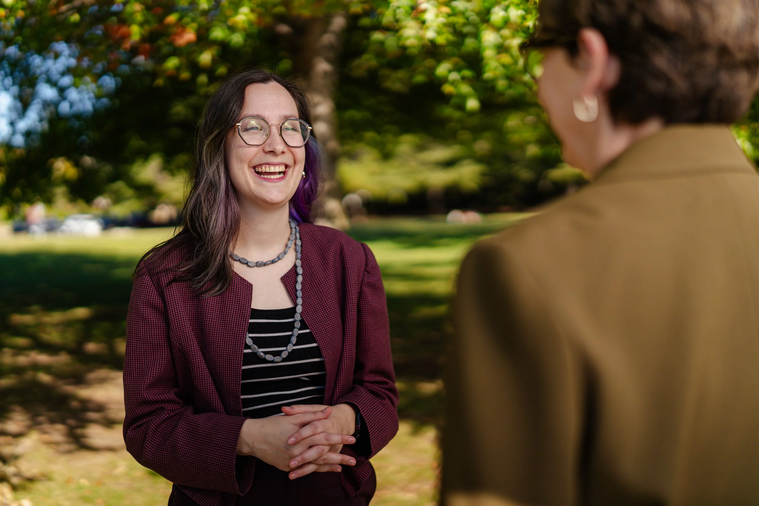 A woman with glasses and purple-tinted hair smiling during a conversation outdoors in a park with green trees in the background.