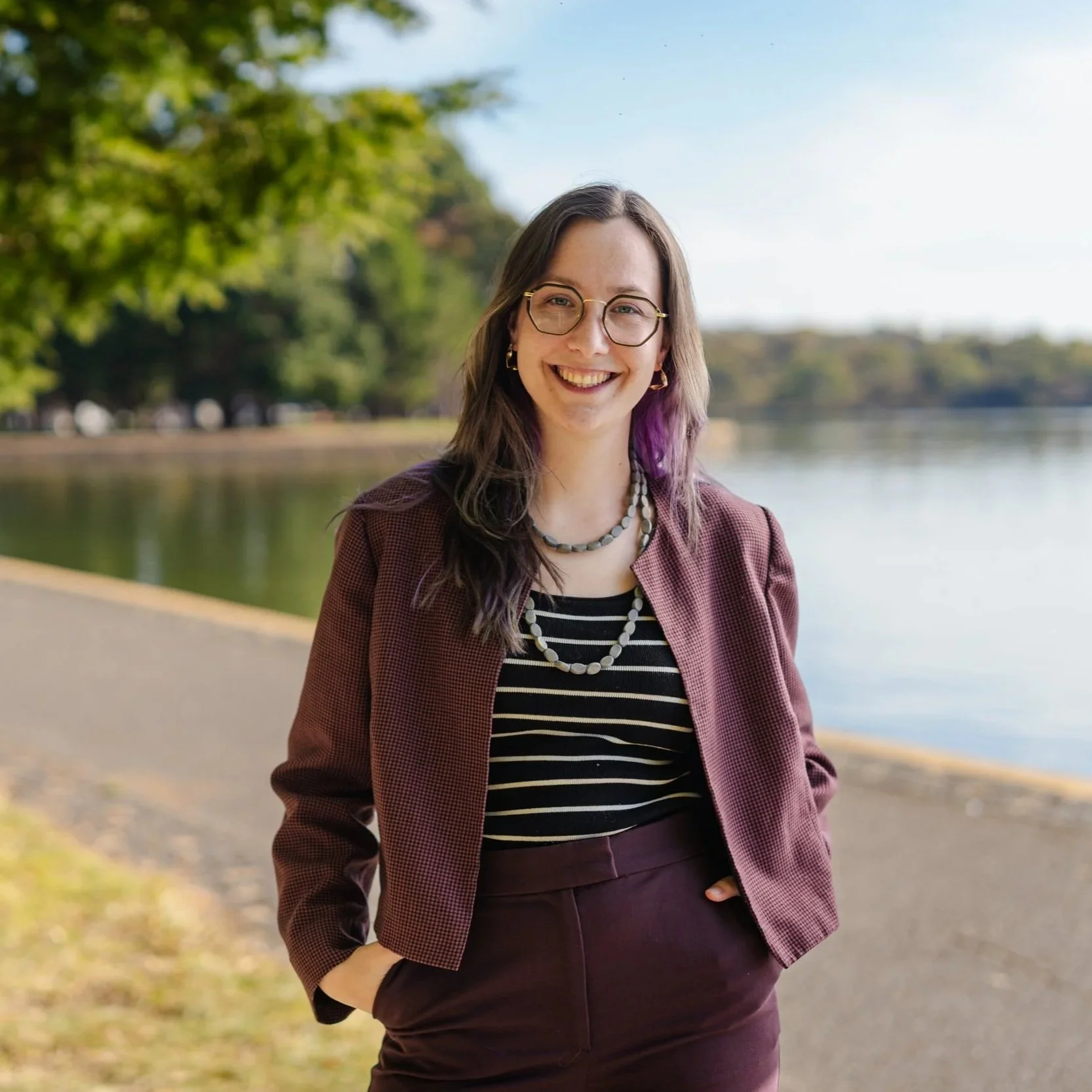 A woman standing outdoors near a body of water, smiling, wearing glasses, a maroon blazer, black and white striped shirt, and maroon pants, with trees and a clear sky in the background.