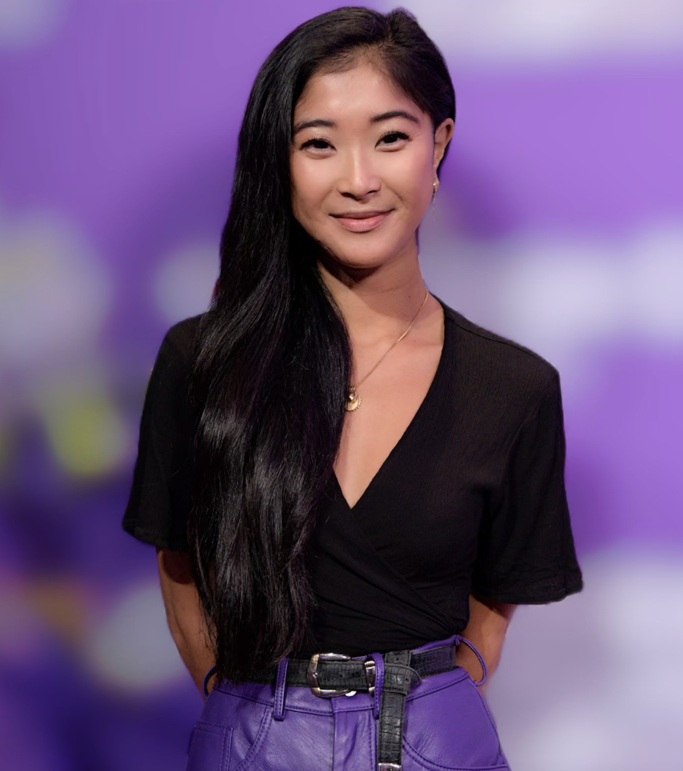 A young woman with long black hair and light skin smiling, standing against a purple and green blurred background. She is wearing a black top, purple pants, and a gold necklace.