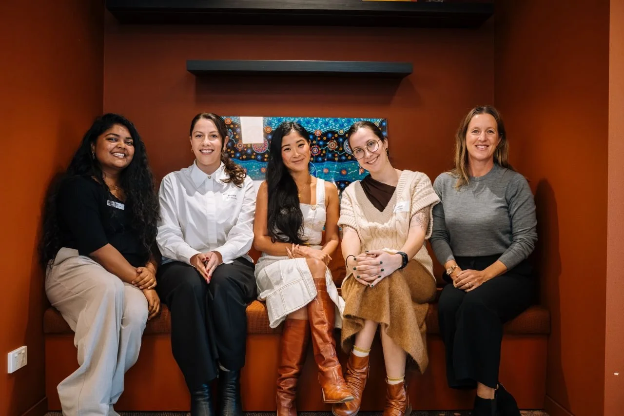 Five women sitting on a bench in a room with a painting on the wall behind them, smiling at the camera.