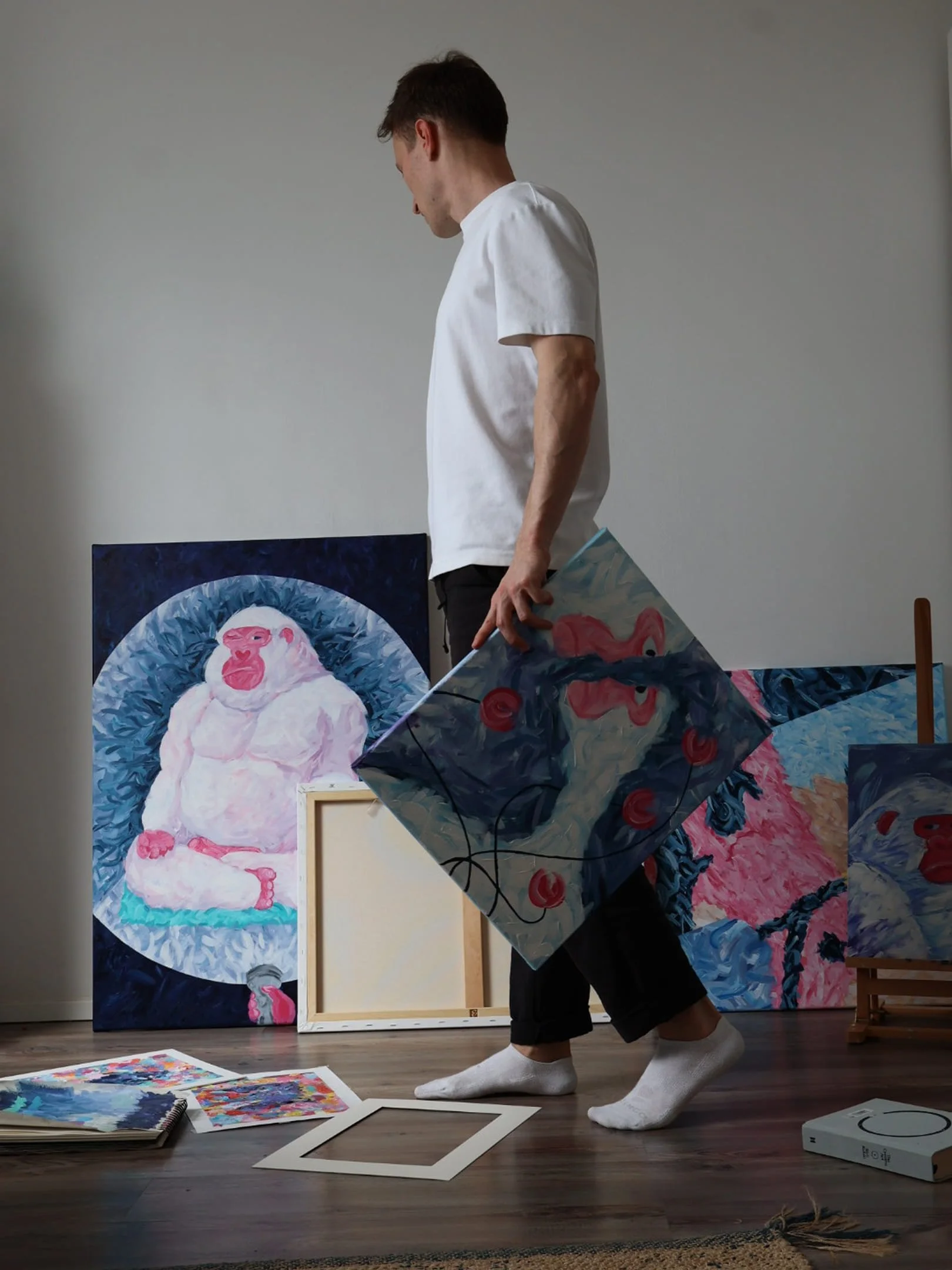 A person kneeling on the floor surrounded by colorful paintings and art supplies in an art studio.