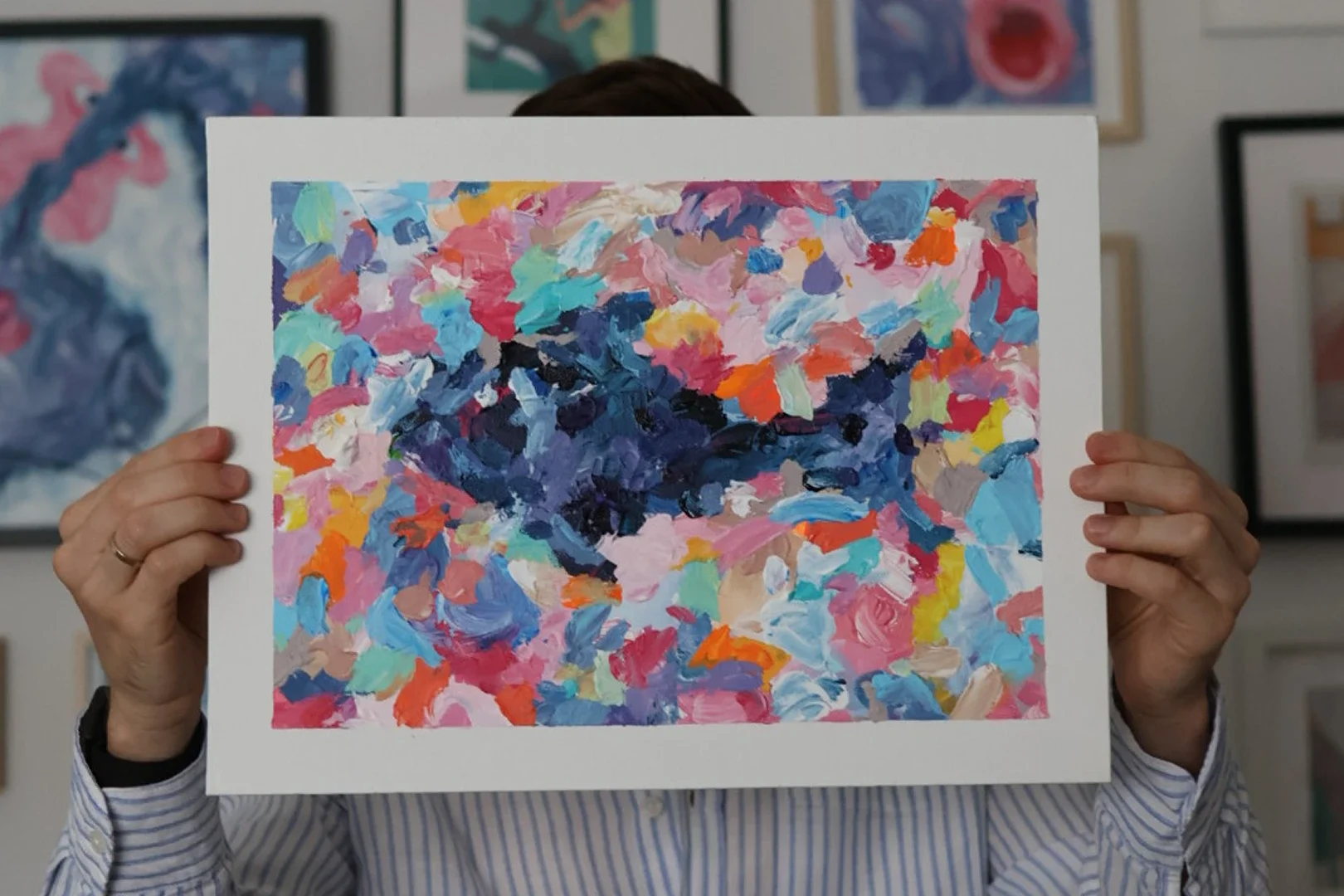Person holding up a colorful abstract painting in front of their face, with other framed artworks visible on the wall behind.