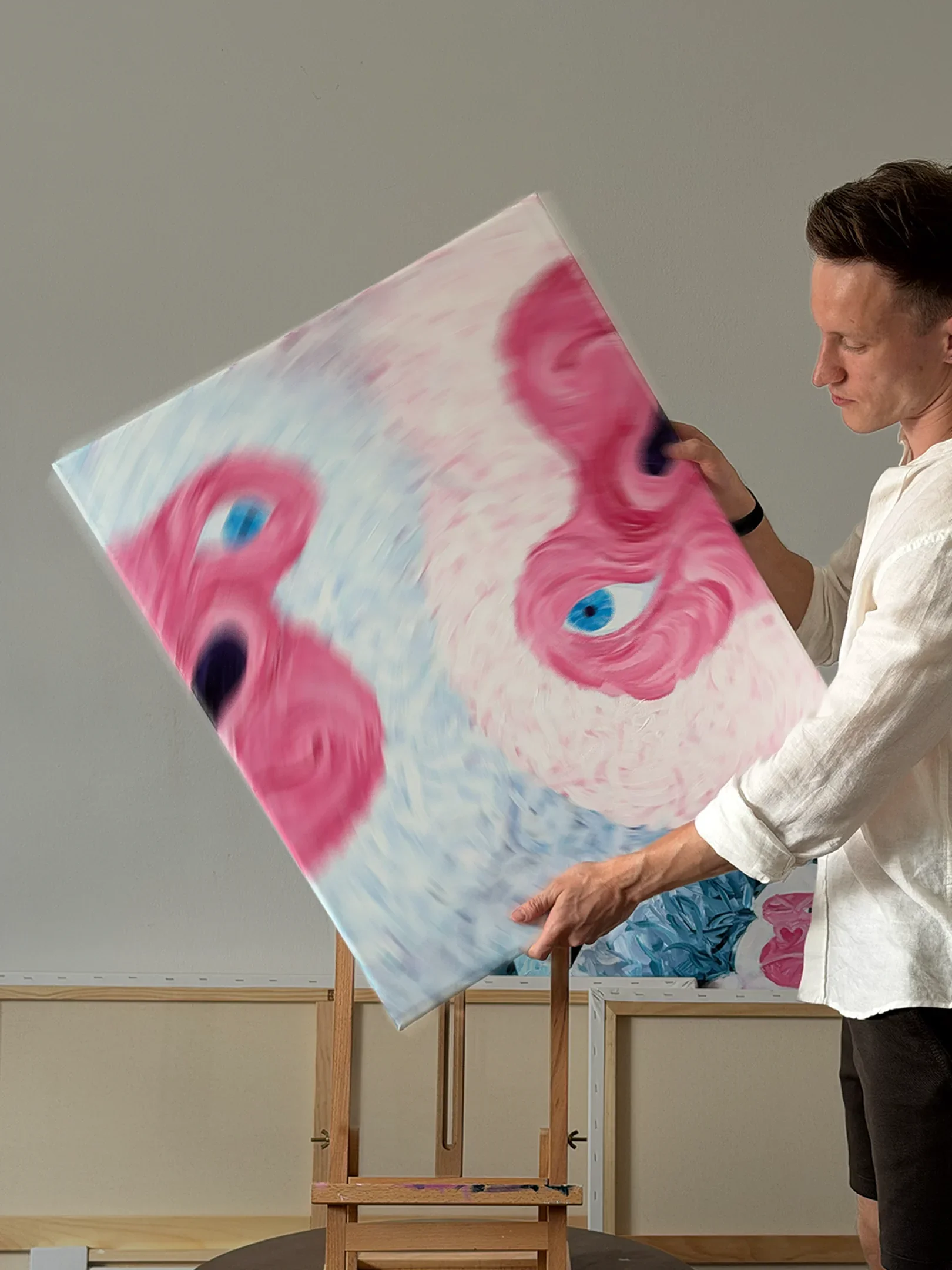 An artist holds a colorful abstract painting with swirling pink, blue, and purple hues, standing in front of an easel and a blank canvas in an art studio.