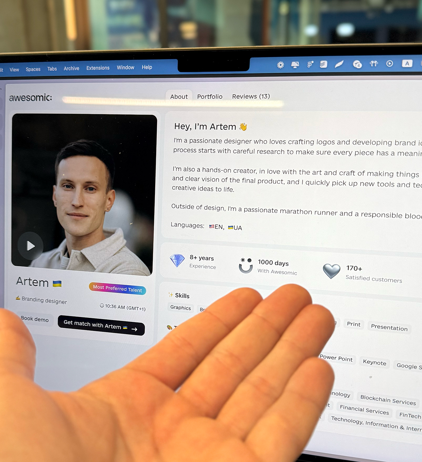A person is holding a tablet displaying a profile of Artem, a branding designer from Ukraine. The profile includes a photo of Artem, a brief introduction, skills, experience, and customer satisfaction metrics.