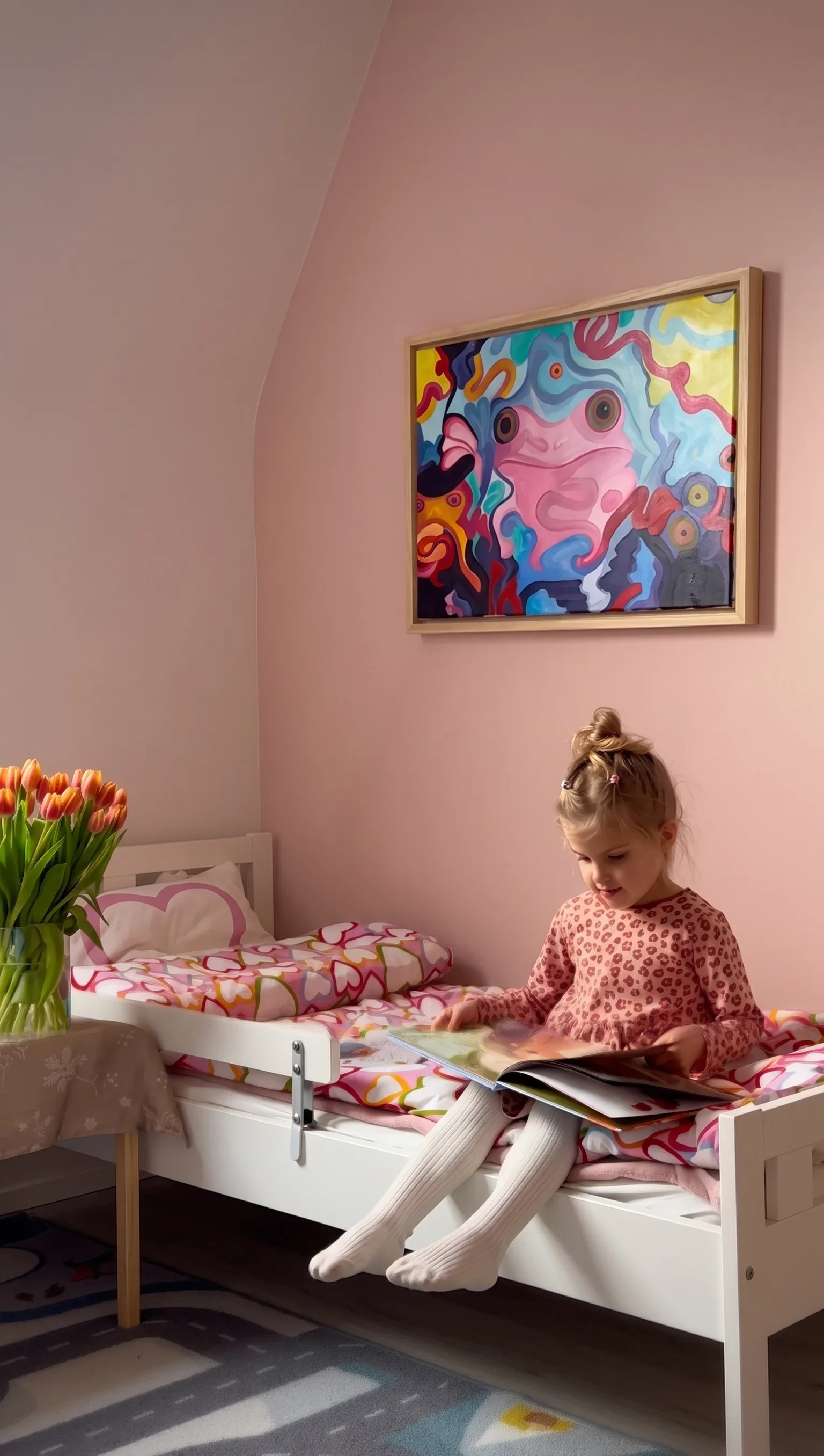 A young girl in a pink and brown leopard print top sitting on a bed with colorful heart-patterned bedding, looking at a book. There is a bouquet of orange tulips on a bedside table and a colorful abstract painting on the pink wall above her.