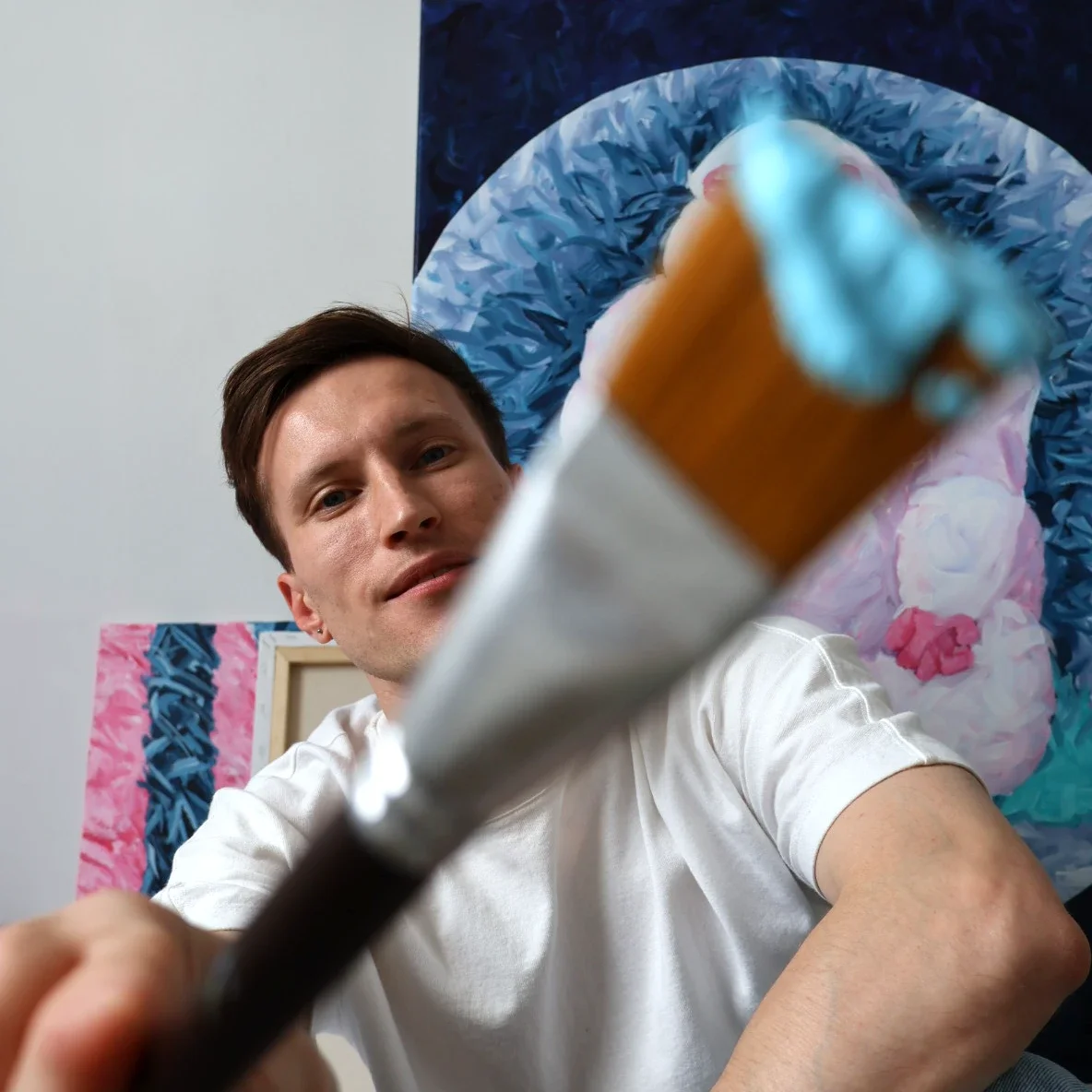Young man with dark hair and earrings holding paintbrush in front of colorful abstract painting.