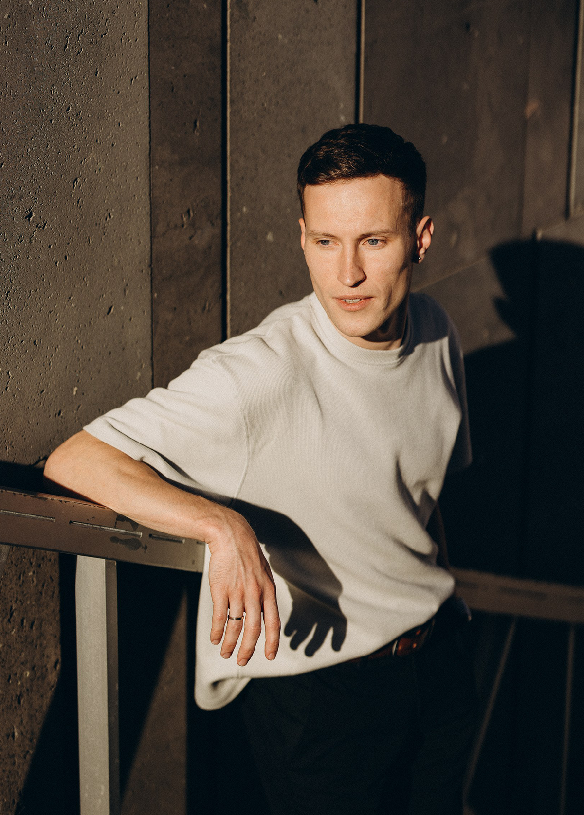 A young man with short dark hair, wearing a light gray T-shirt, sitting against a dark textured wall.