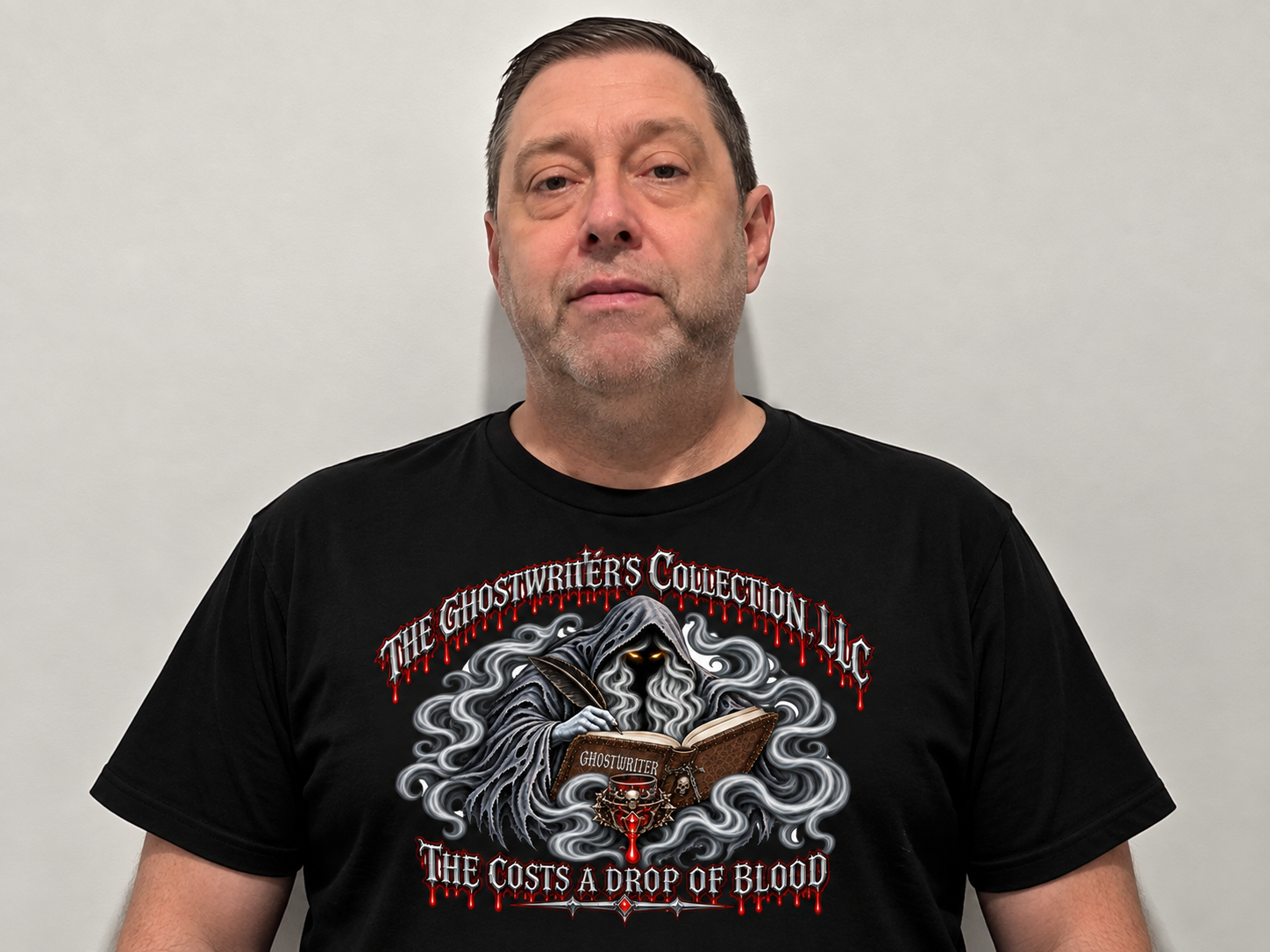 A man with short dark hair and stubble, wearing a black T-shirt with a gothic design of a hooded ghostly figure writing in a book, surrounded by smoke, with red and white text that reads 'The Ghostwriter's Collection LLC' and 'The Cost's a Drop of Blood'.