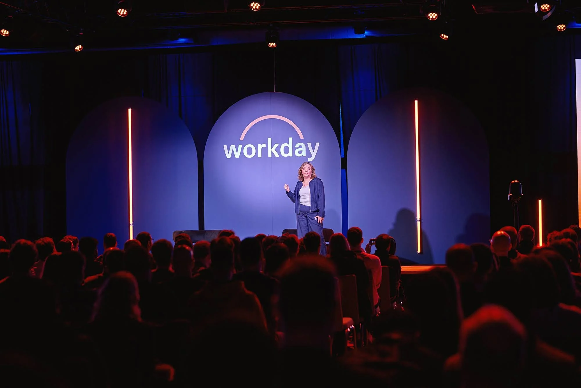 A woman giving a presentation on stage at a conference called Workday, with an audience seated in front of her, dark background, blue and orange lighting.