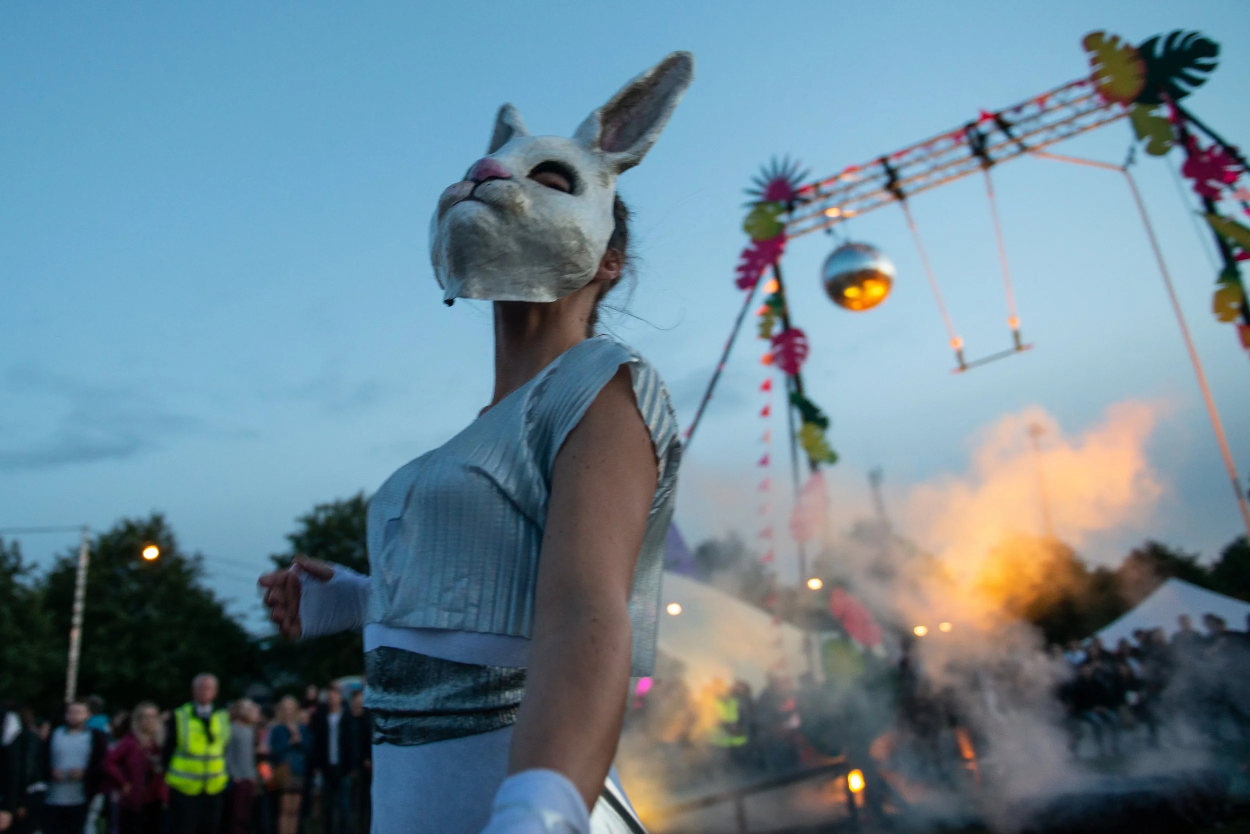 Person wearing a white rabbit mask at an outdoor festival during sunset, with colorful decorations and a crowd in the background.