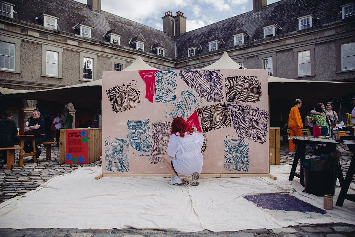 A person with red hair, dressed in white, crouching in front of a large canvas with abstract black, blue, and red paint swirls, in an outdoor courtyard with tents and people.