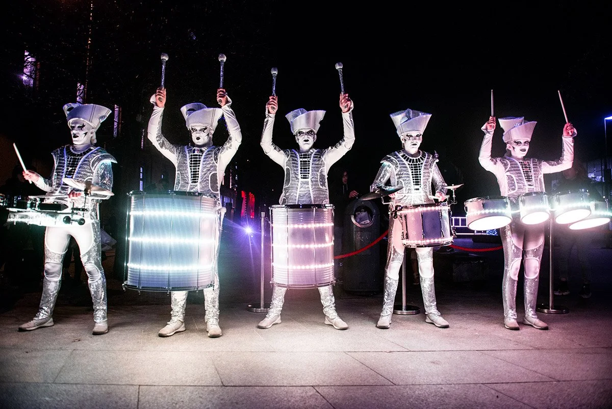 Performers dressed in silver futuristic costumes with LED lights, wearing white masks that resemble porcelain, playing drums and other percussion instruments at a night event.