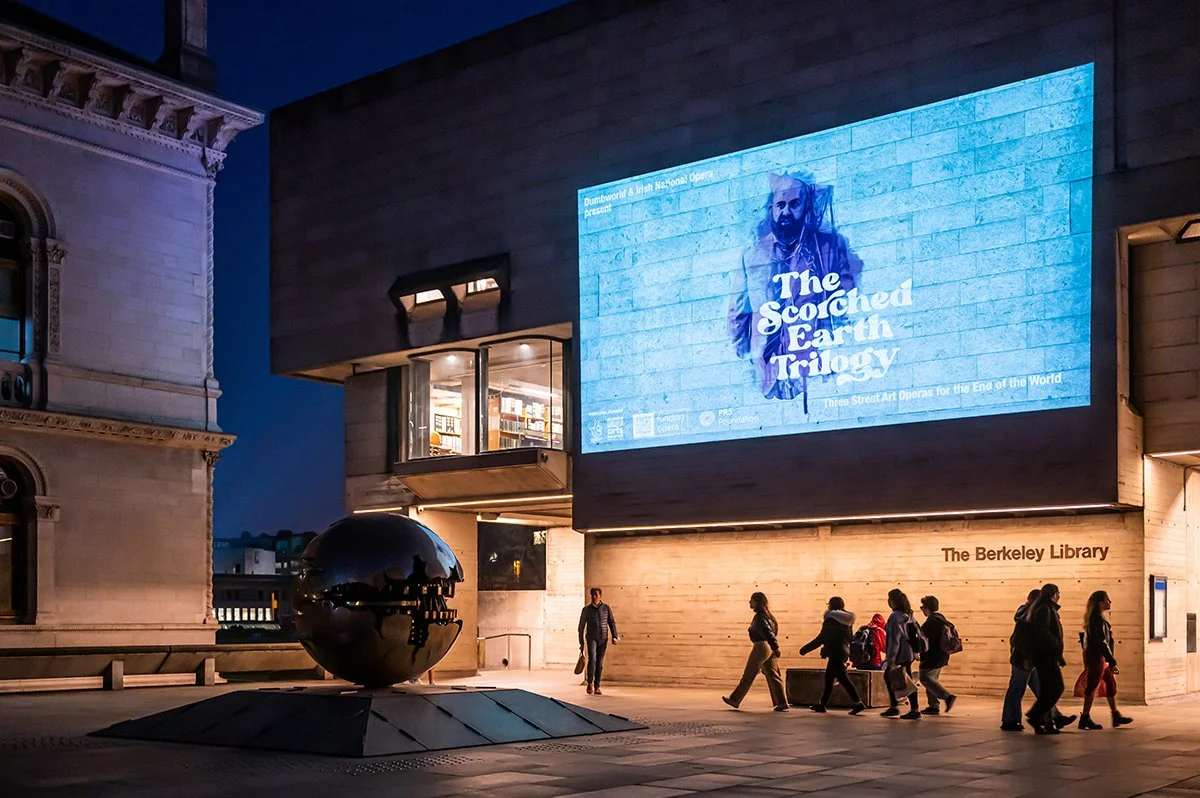 People walking past the Berkeley Library at night with a large digital billboard displaying promotional content for a theatrical production called 'The Scorched Earth Trilogy' on the building's exterior.
