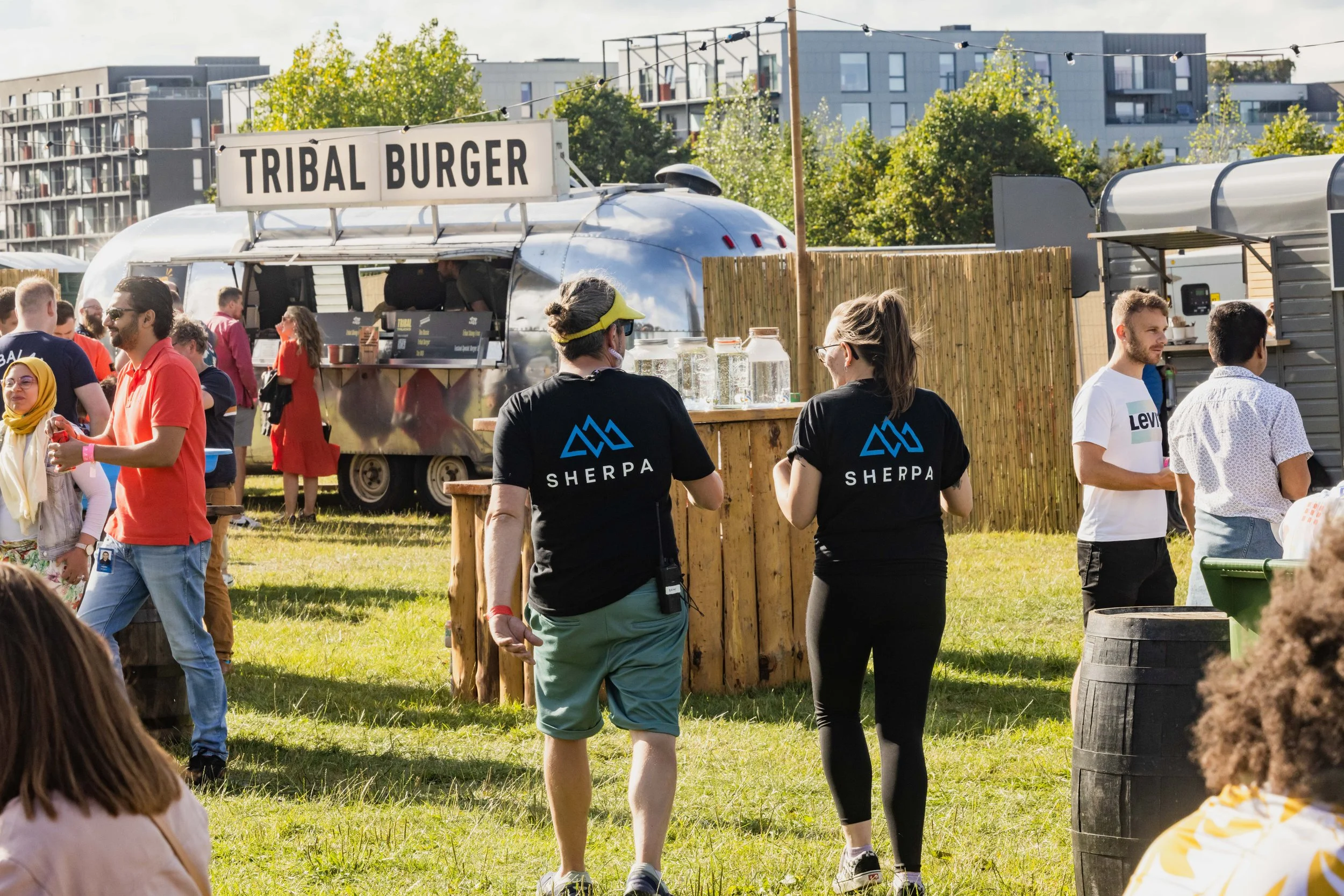 People at an outdoor event near food trucks, with a sign reading 'Tribal Burger', and others engaging in conversations, some wearing black T-shirts with 'SHERPA' logo. The setting has a grassy area, trees, and modern buildings in the background.