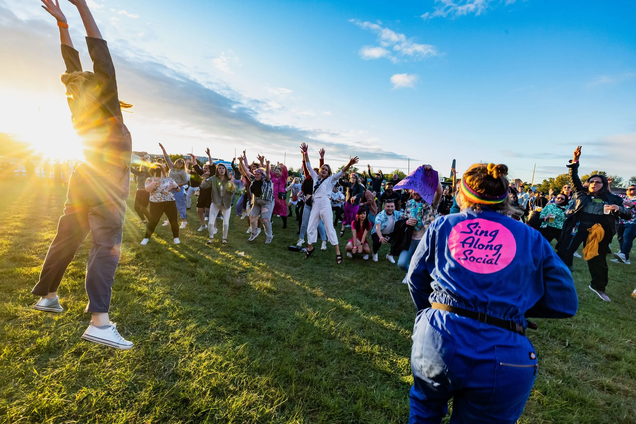 A large crowd of people gathered outdoors during sunset, dancing and celebrating. Many individuals have their arms raised, and some are wearing colorful clothing and accessories. The scene exudes a festive, joyful atmosphere.